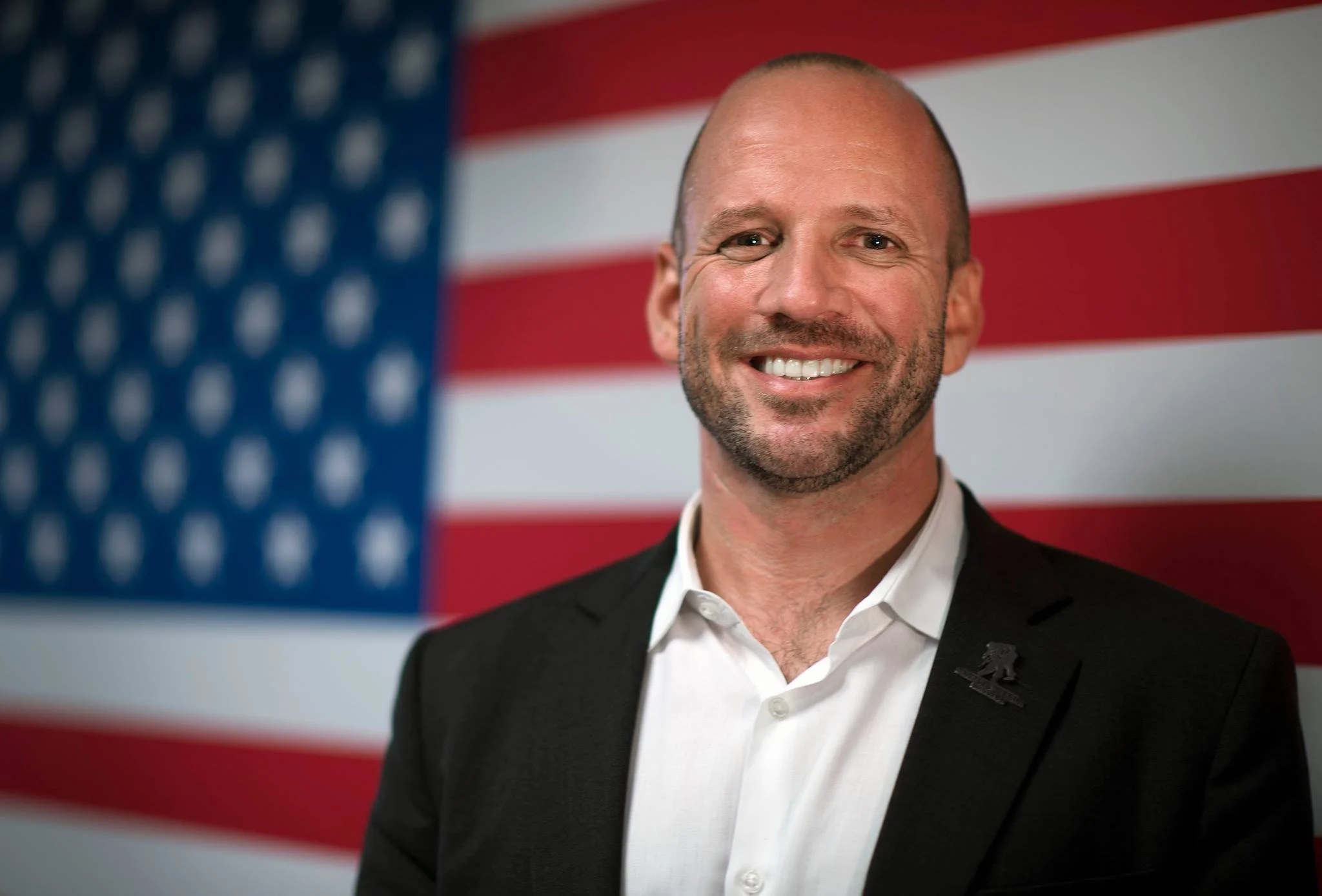 Dan Nevins is in a black suit and white shirt, standing in front of the American flag.