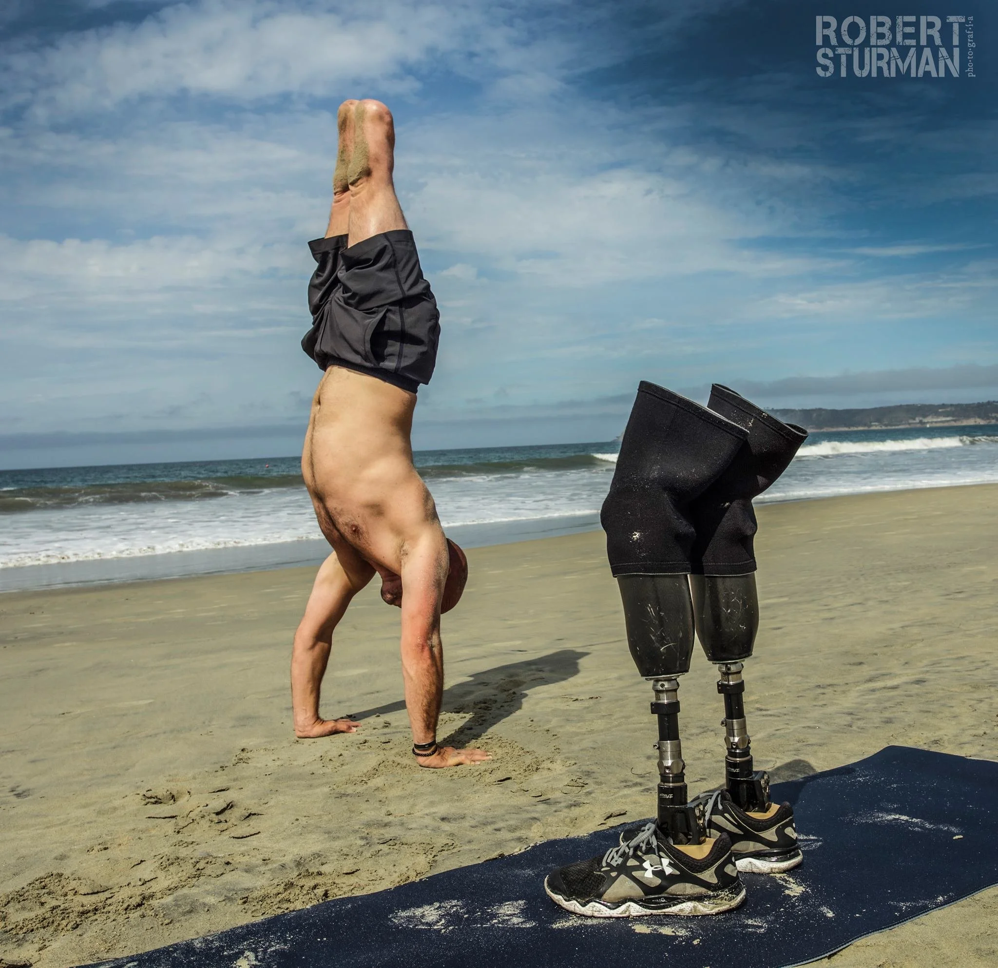 Dan Nevins performing a handstand on the beach, with his legs extended upward. Next to him is a pair of black prosthetic legs positioned on a mat, with a pair of athletic shoes at the base.