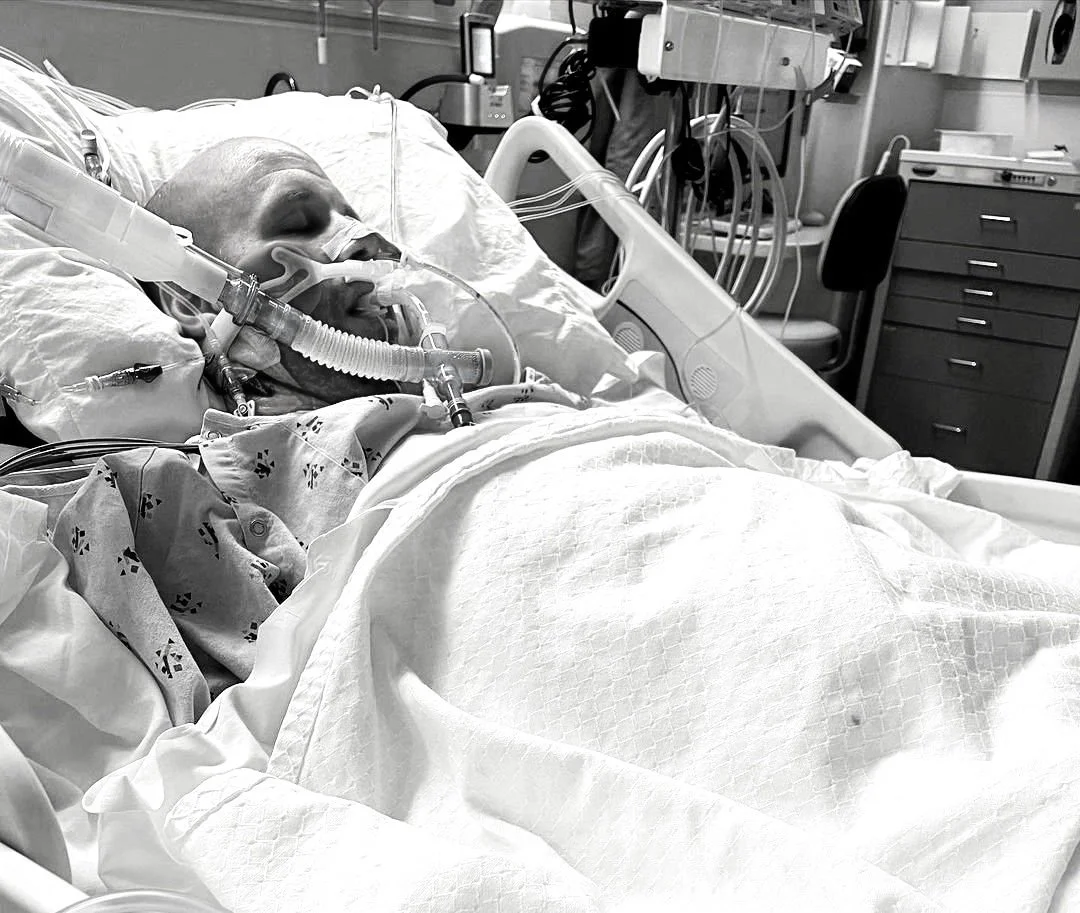 Dan Nevins laying in a hospital bed with a ventilator and various medical tubes and wires connected to him.