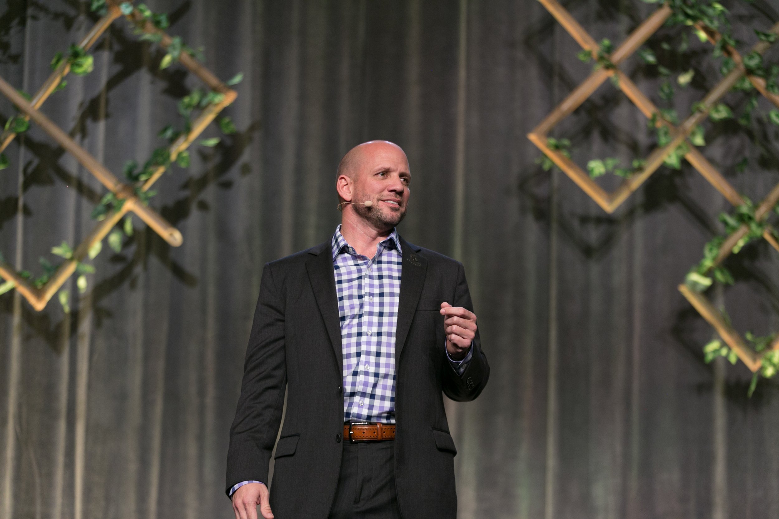 A man in a black suit and checkered shirt speaking on stage with a headset microphone, against a backdrop of gray curtains and decorative wooden lattice with green leaves.