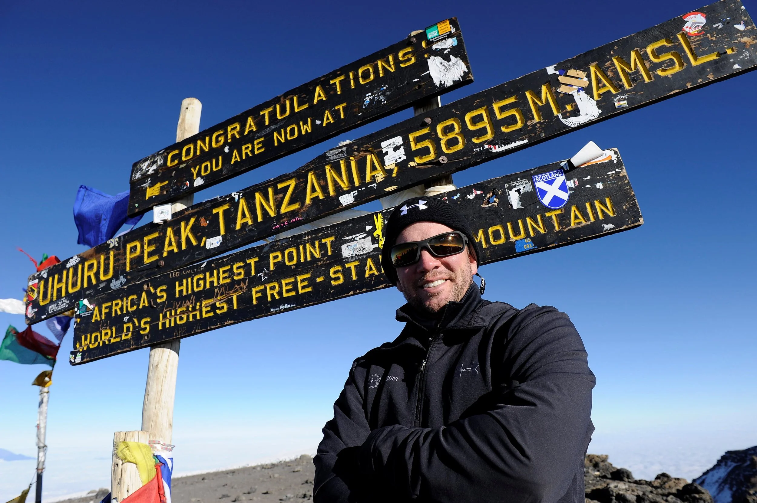 Dan Nevins standing in front of a wooden sign at Uhuru Peak on Mount Kilimanjaro, Tanzania, indicating the highest point in Africa and the world’s highest free-standing mountain.