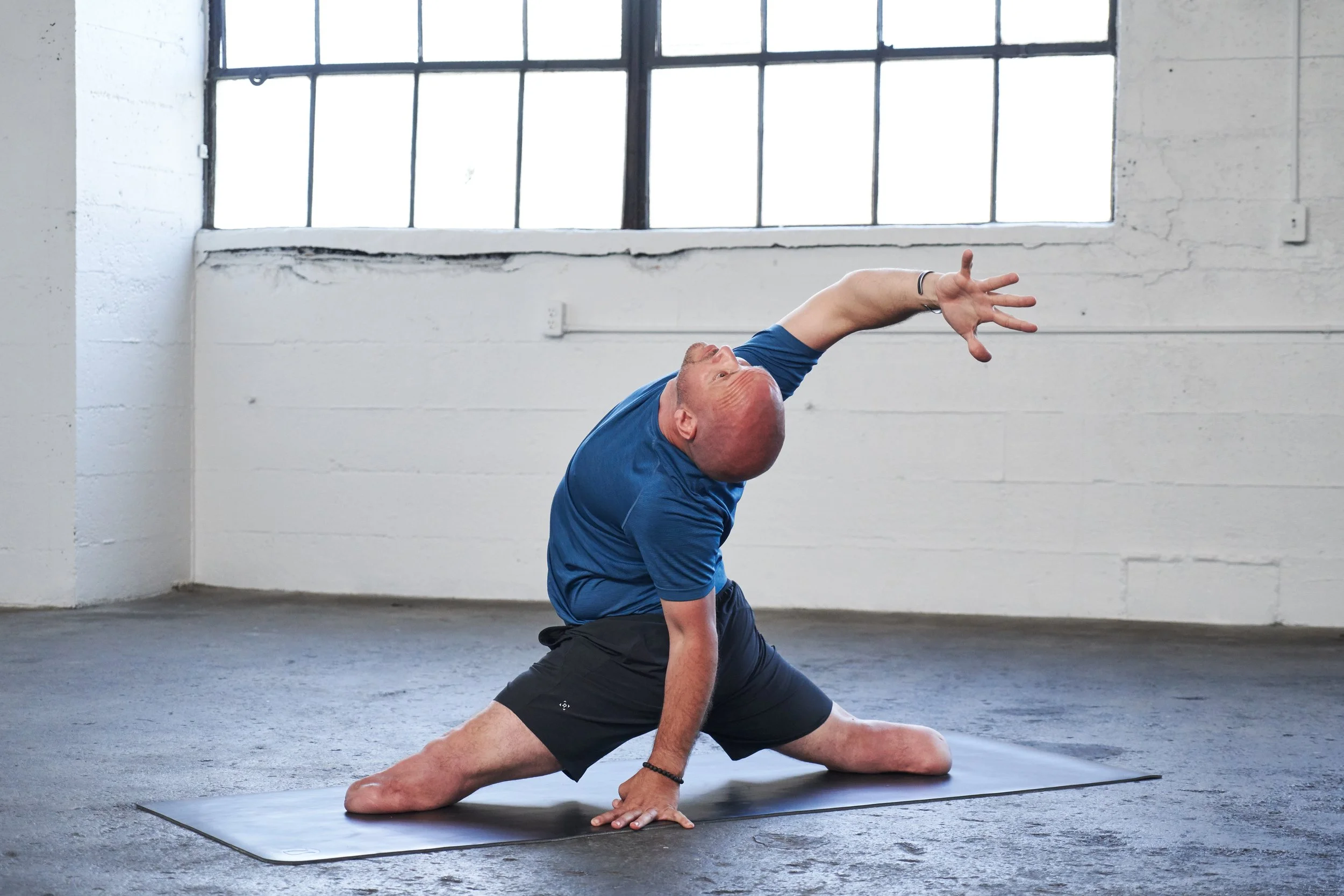 Dan Nevins performing a yoga pose on a black yoga mat in a bright, industrial-style room with large windows.