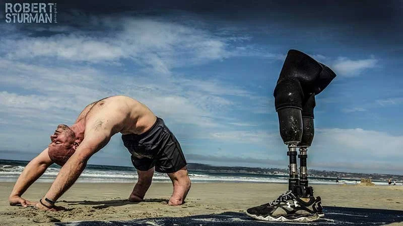 Dan Nevins with a prosthetic leg doing a backbend on the beach, with the prosthetic leg on the sand, wearing athletic shoes, under a partly cloudy sky.