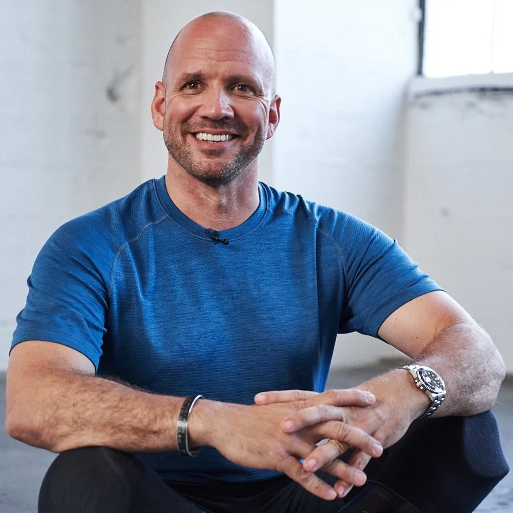 Dan Nevins is smiling, wearing a blue athletic shirt, sitting with his hands clasped. He is indoors near a window.