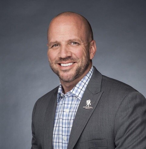 A professional headshot of Dan Nevins wearing a gray suit jacket and a checked shirt, against a gray background.