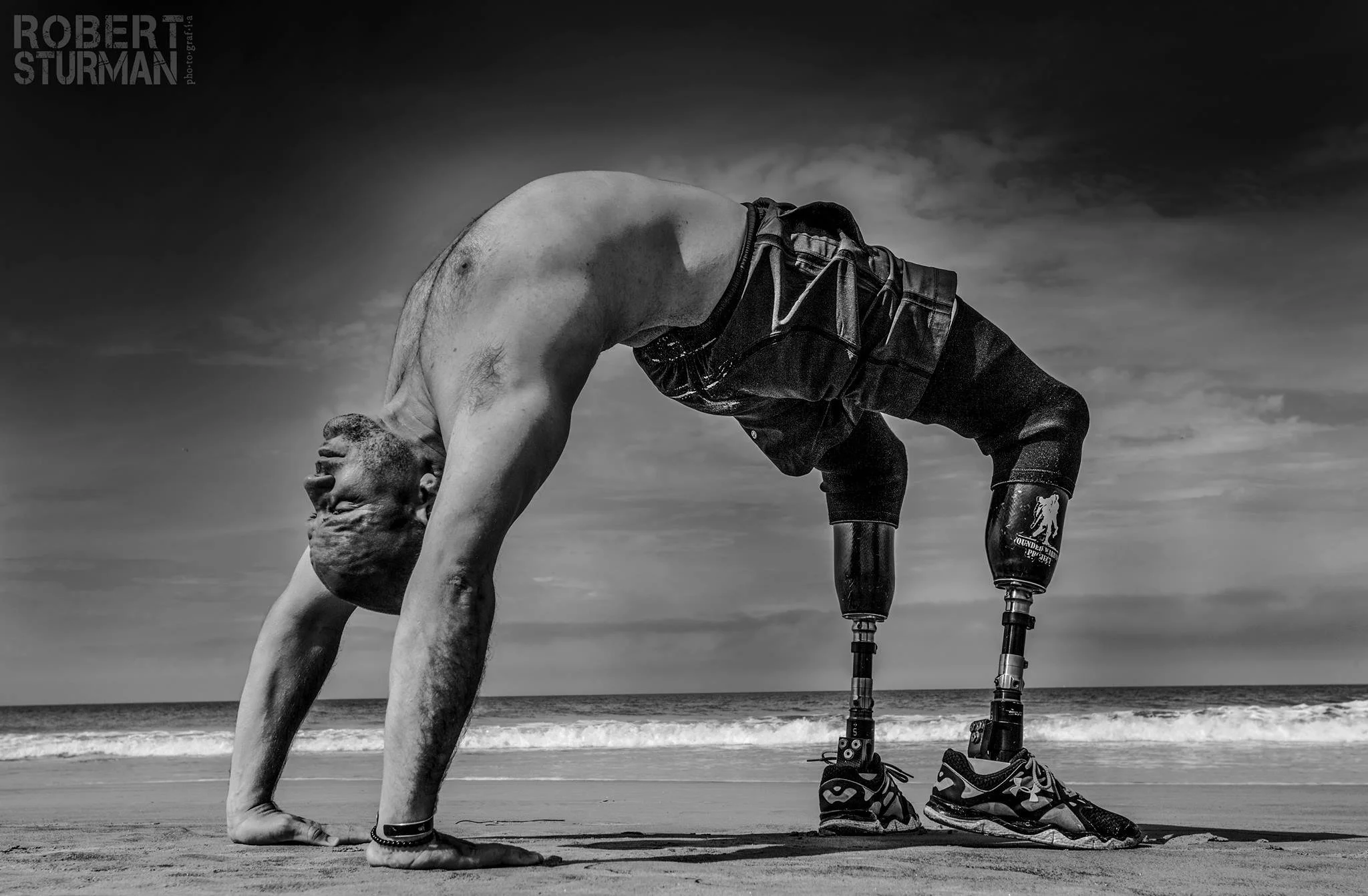 Dan Nevins does a backbend with prosthetic legs on a beach, with the ocean and sky in the background.