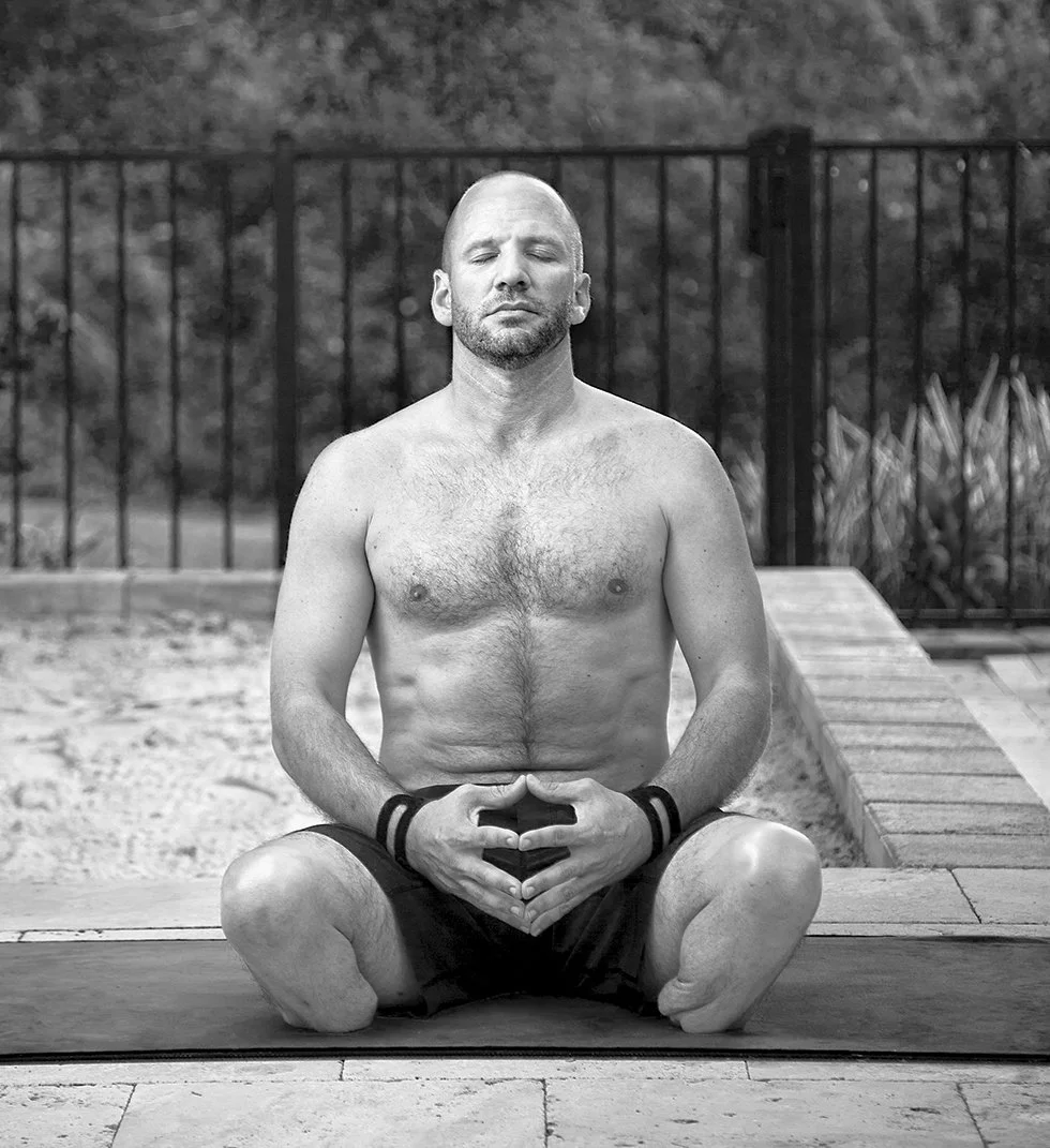 Dan Nevins practicing meditation outdoors, sitting cross-legged on a yoga mat in front of a fence with a garden background, eyes closed, appearing peaceful.