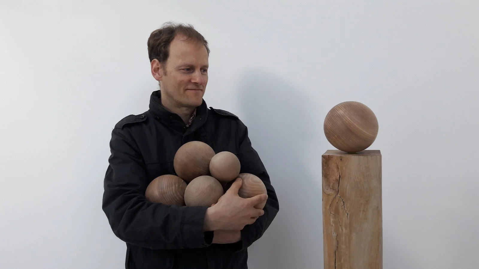 Man holding several wooden spheres, standing next to a wooden pedestal with a single wooden sphere on top.