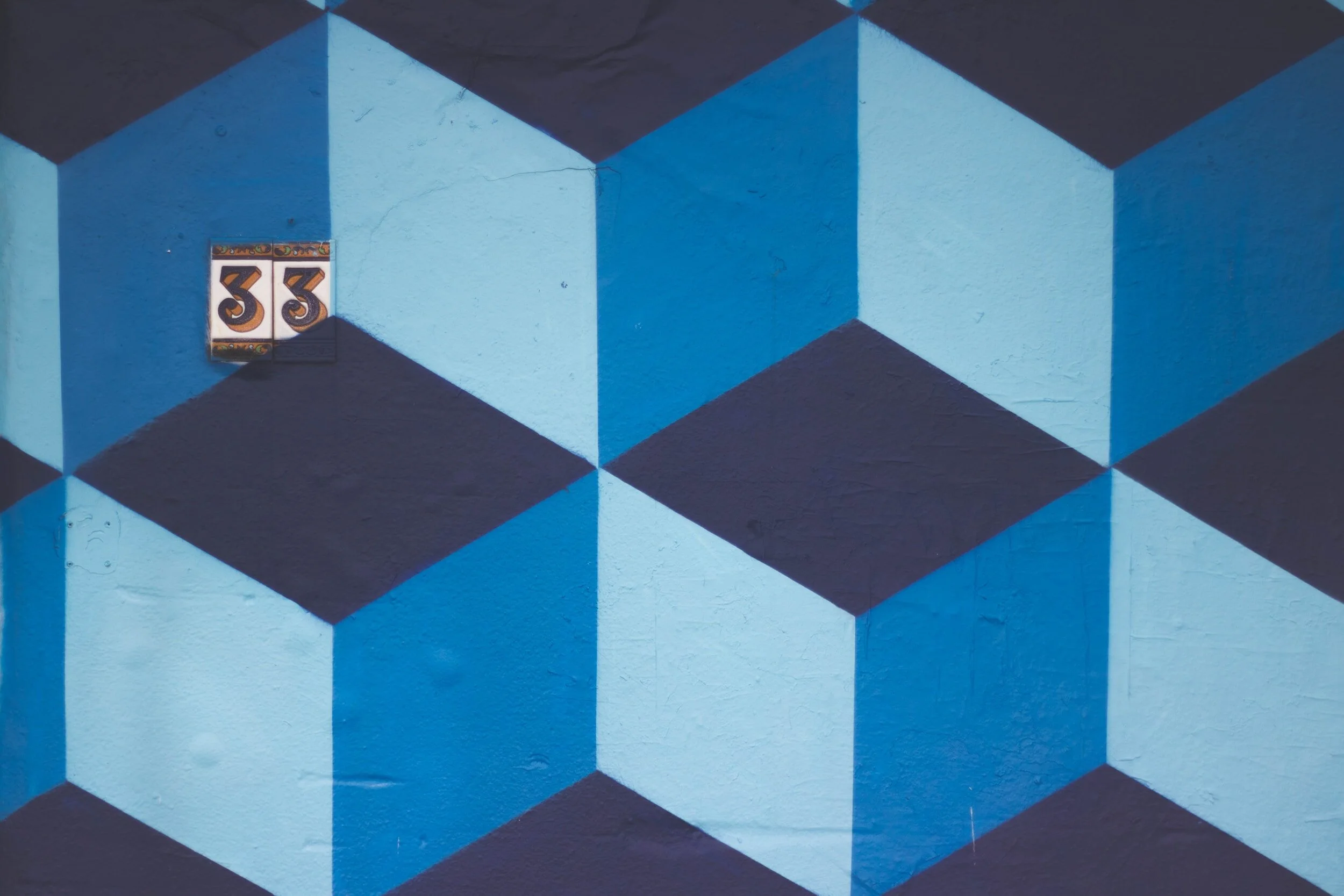 A geometric mural painted on a wall with blue, black, and white shapes creating a 3D cube pattern. In the upper left corner, there is a tile with the number 33.