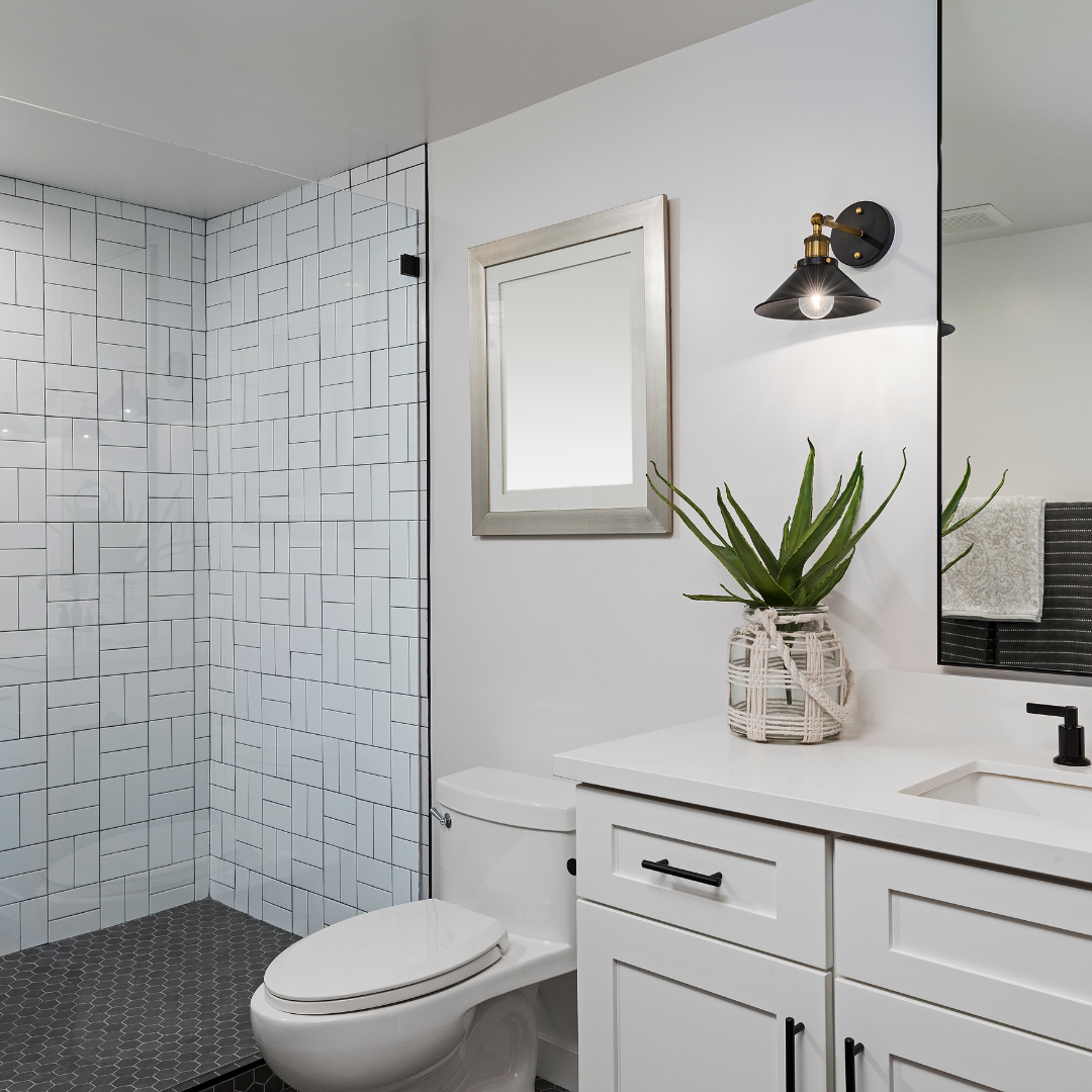 Modern bathroom with a walk-in shower featuring white tiles and black grout, a white toilet, a white vanity with black handles, a potted plant on the vanity, a mirror, and a wall-mounted black and gold light fixture.