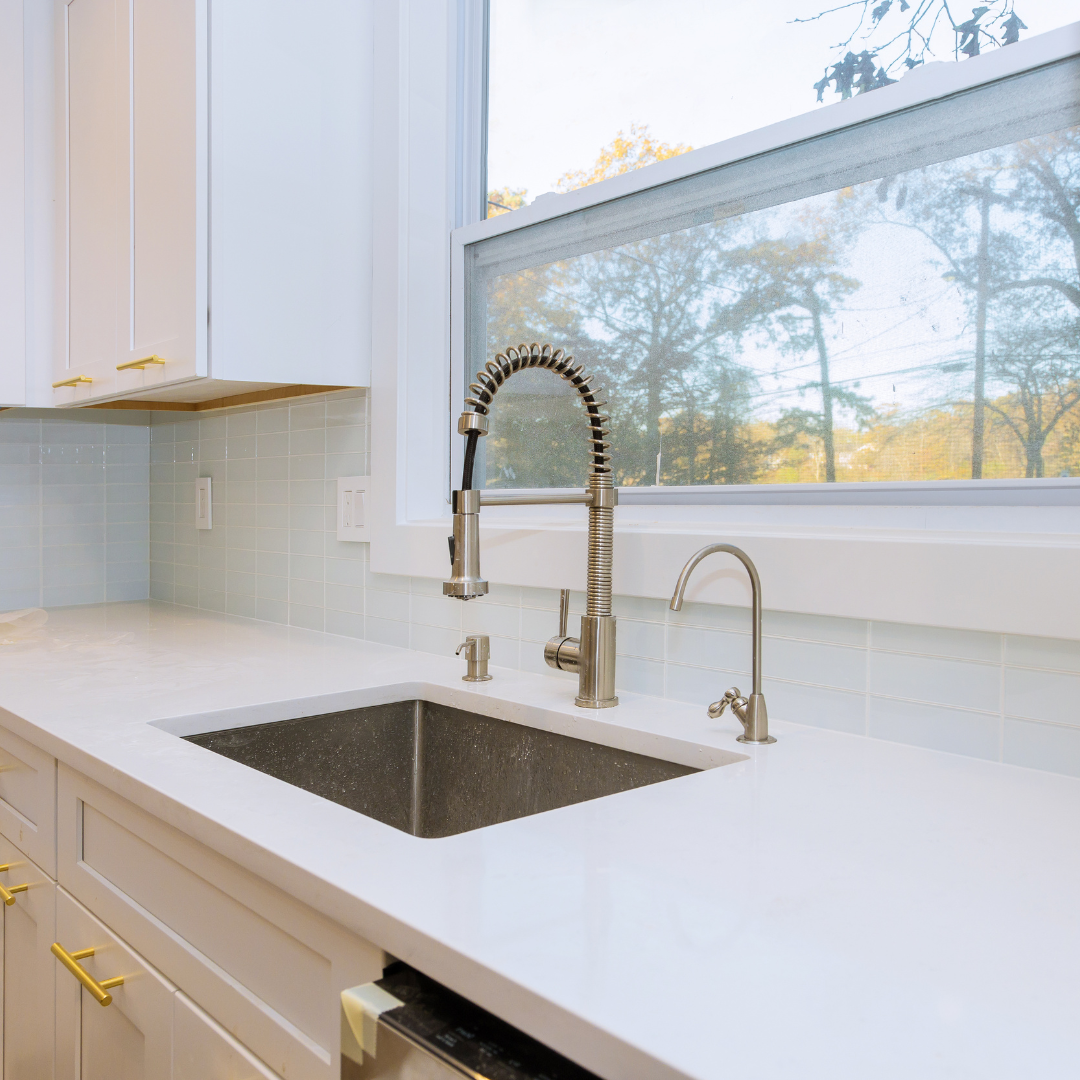 Modern kitchen sink area with a large window showing outdoor trees, stainless steel faucets, white countertops, and light-colored cabinets.