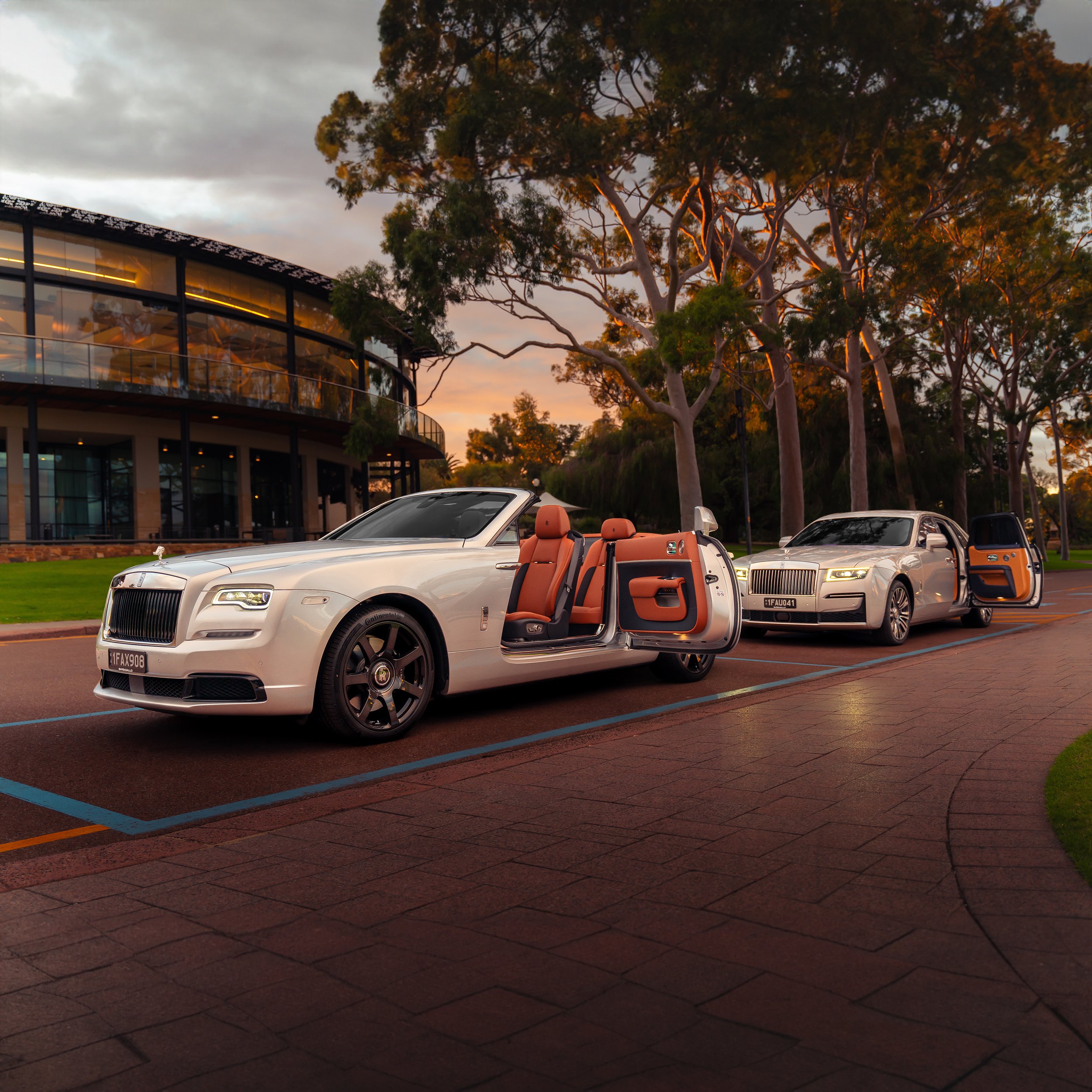 Luxury white cars with open doors parked on a street, trees and a modern building in the background.