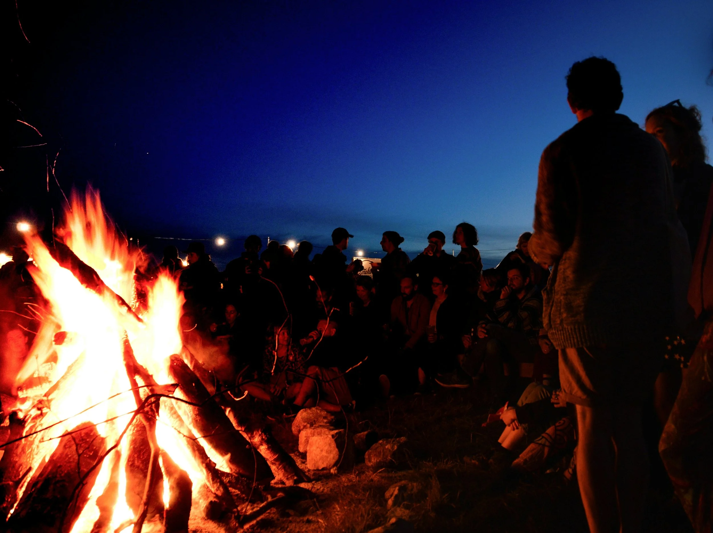A group of people gathered around a large campfire at dusk, silhouetted against a deep blue night sky