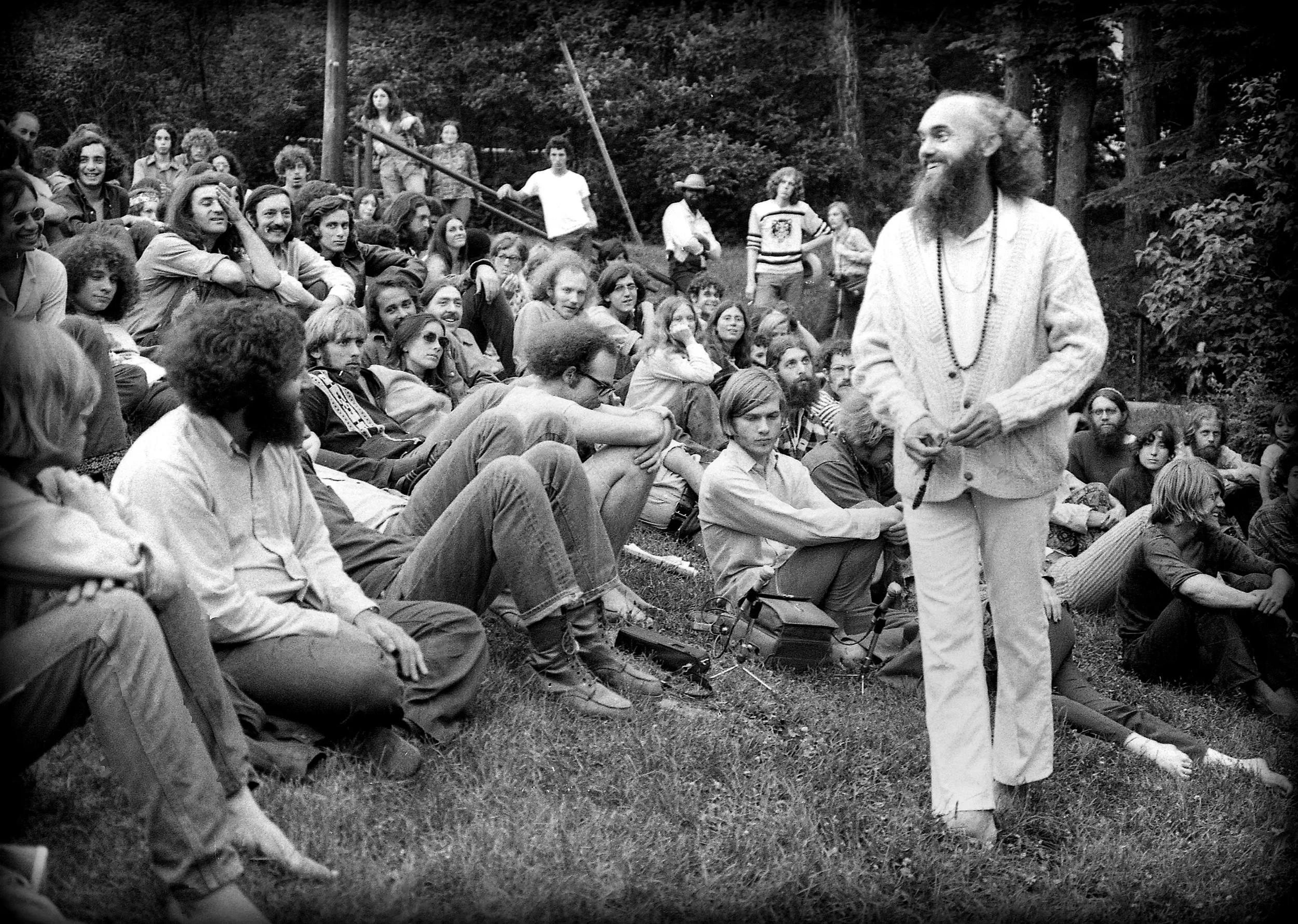 Ram Dass speaking to a crowd of young people seated on grass outdoors, 1970s