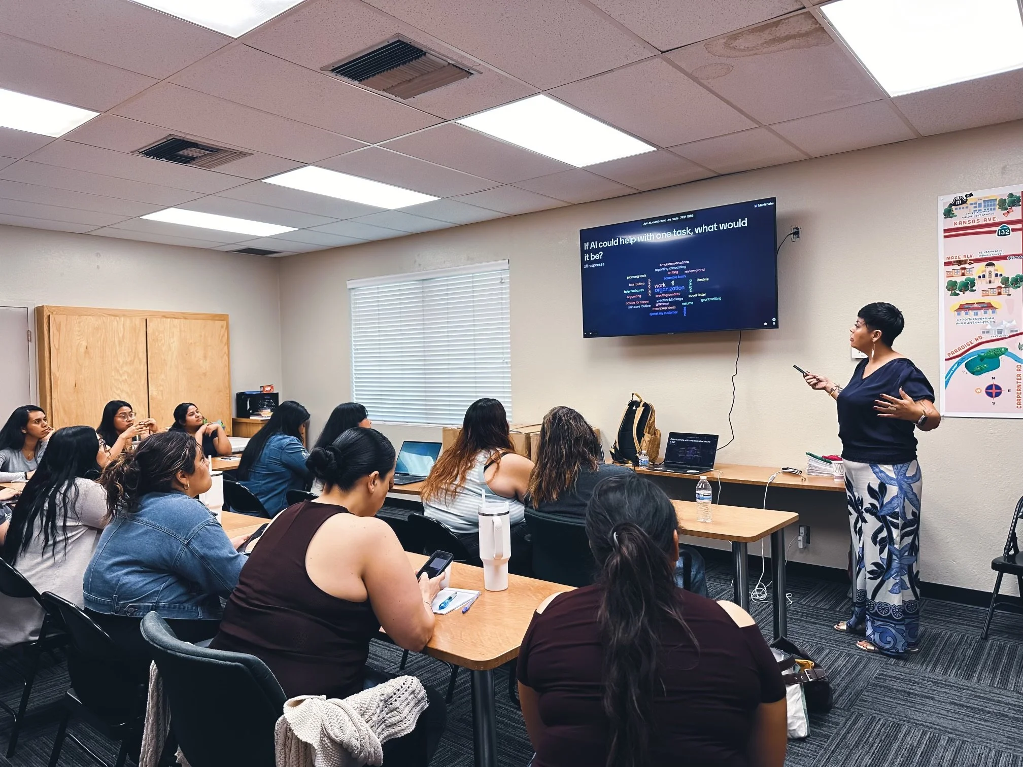 Classroom with a teacher standing at the front presenting to a group of students seated at desks, viewing a screen with a presentation. Students are listening attentively and taking notes. The room has overhead lighting and educational posters on the walls.