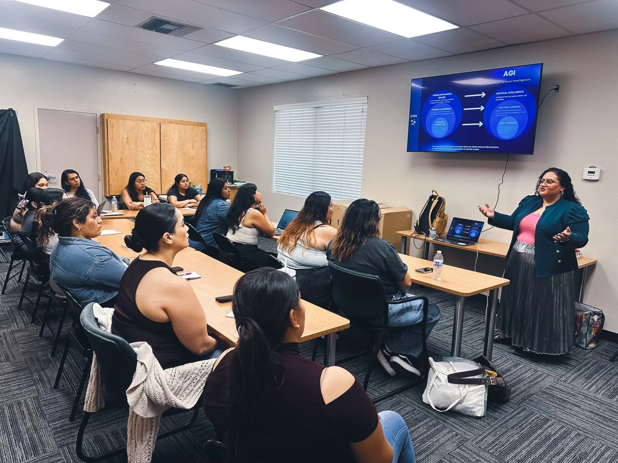 A woman is giving a presentation to a group of women seated at tables in a classroom setting. A screen displays a slide titled "AGI" with diagrams and text. The attendees are focused and taking notes. The room has a modern, professional appearance with carpeted flooring and fluorescent lighting.