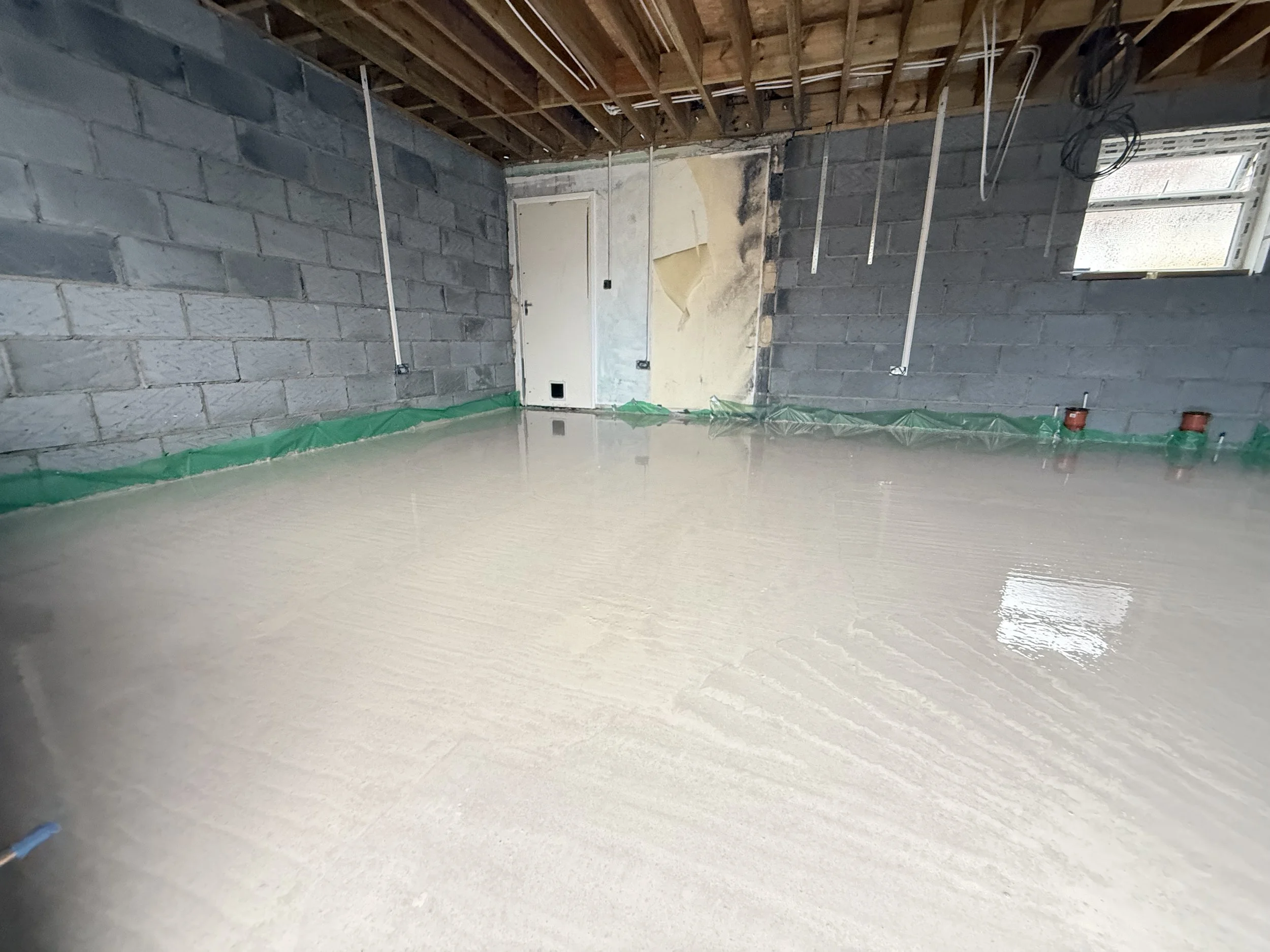 Empty room under renovation with bare cinder block walls, exposed wooden ceiling beams, and a shiny new concrete floor, with electrical wiring hanging from the ceiling and small windows near the ceiling.