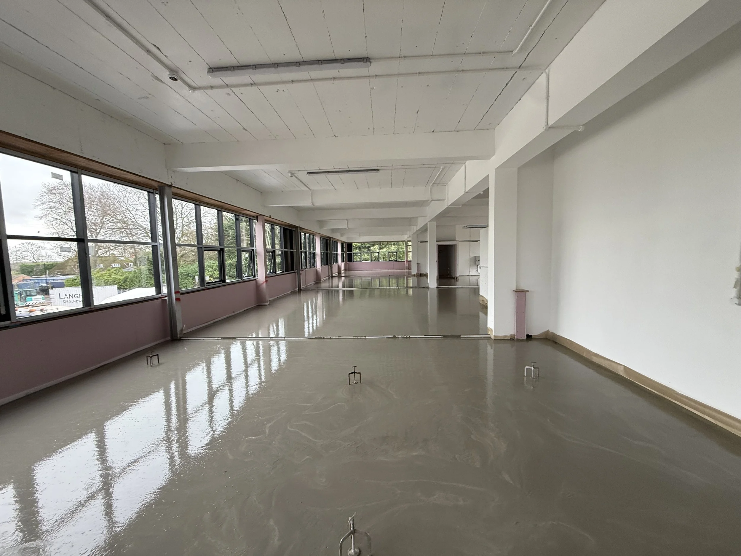 An empty room with large windows along one wall, a polished concrete floor, and a white ceiling with exposed pipes and lighting fixtures.