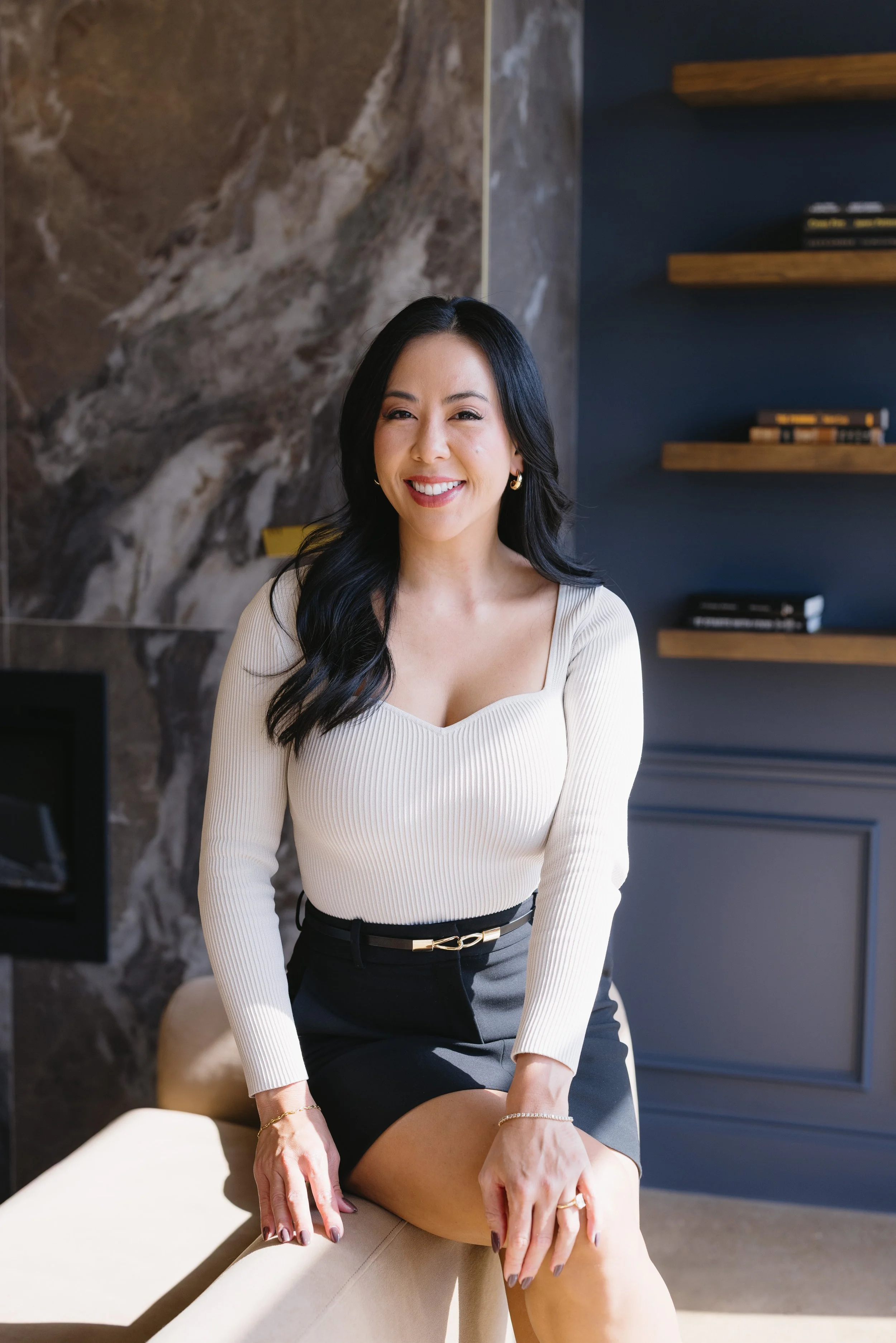 Portrait of a smiling woman with long black hair, wearing a cream-colored ribbed top and black skirt, sitting on an armrest in a modern room with marble and blue wall decor.