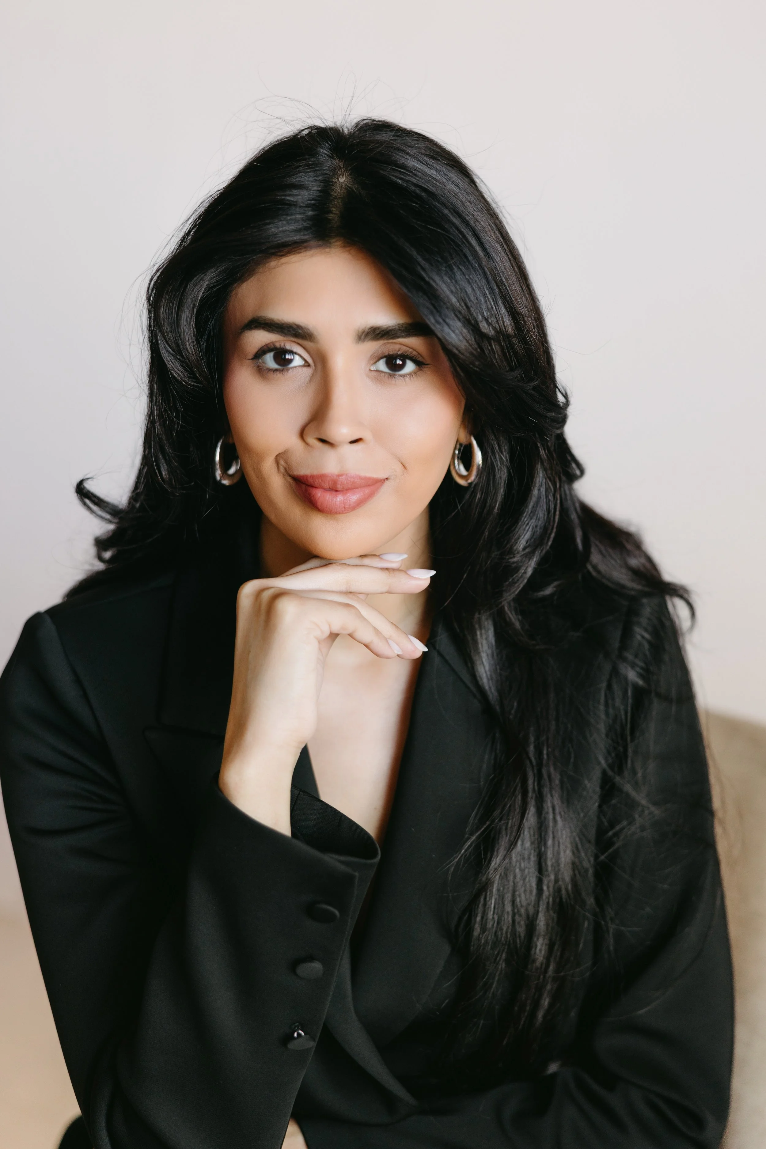 Portrait of a smiling woman with long black hair, wearing a cream-colored ribbed top and black skirt, sitting on an armrest in a modern room with marble and blue wall decor.
