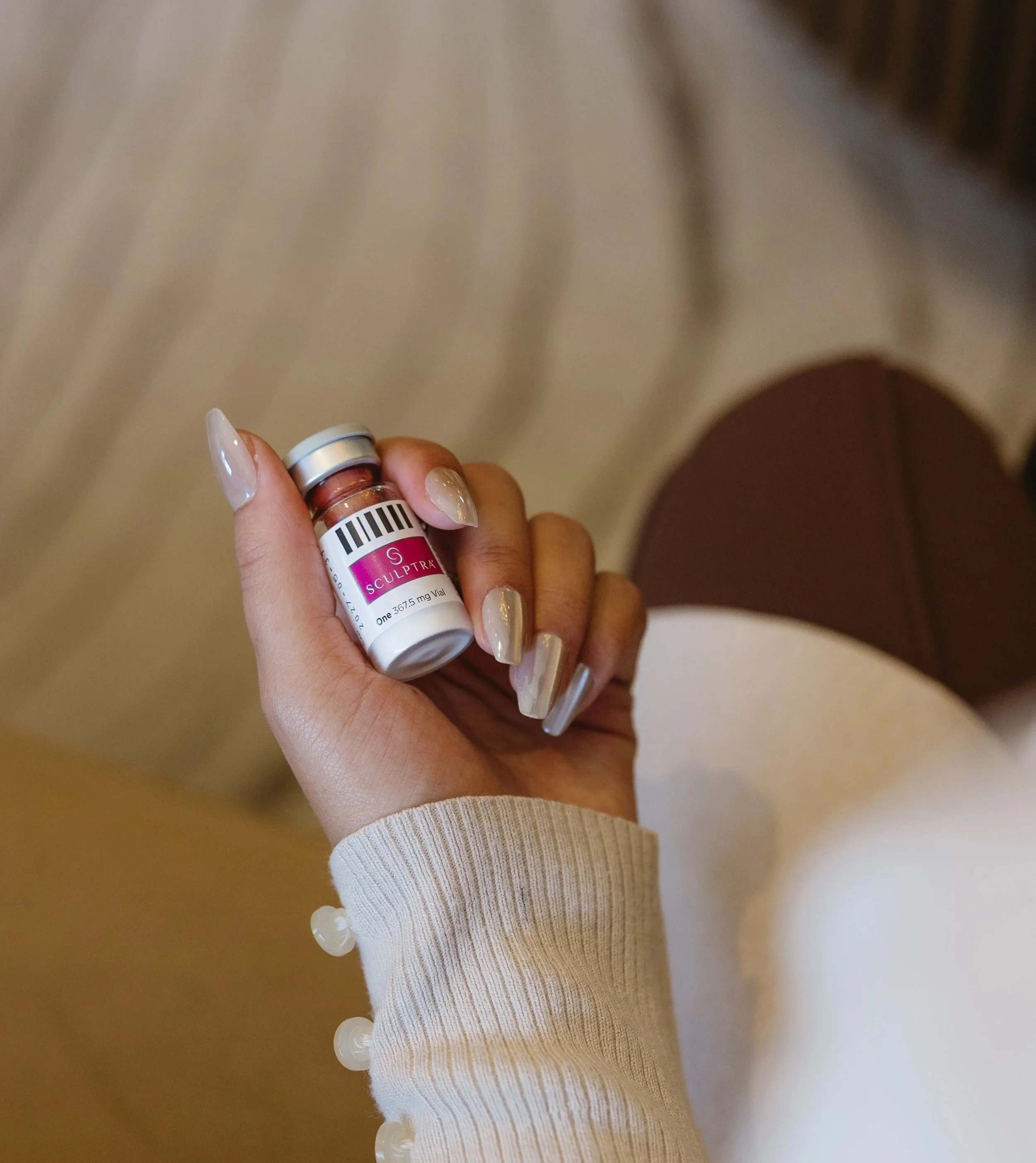 A hand with manicured nails holds a small bottle of Sculptra, a cosmetic injectable. The person is wearing a beige sweater with decorative buttons on the sleeve.