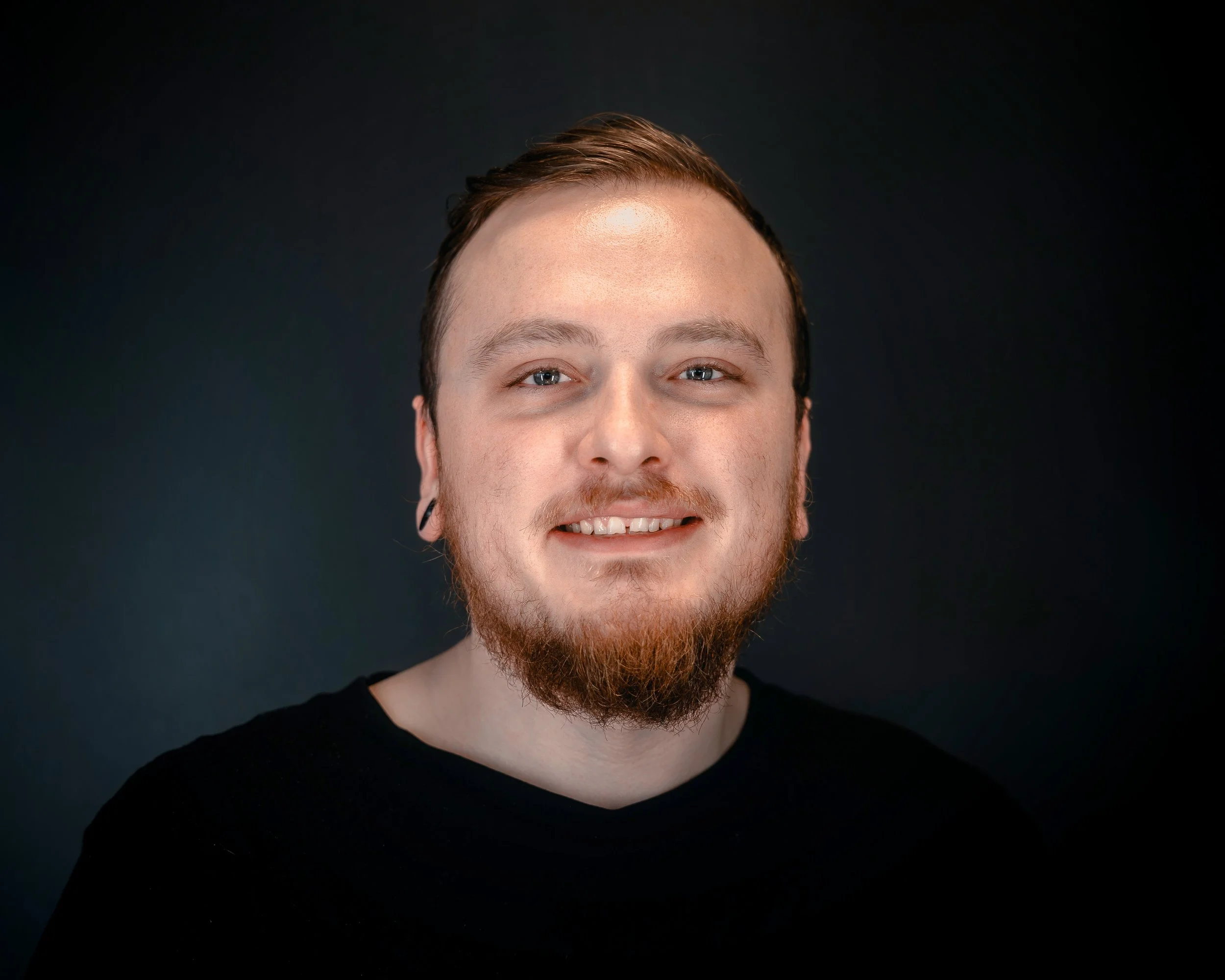 Smiling person with tattoos and gauged ears wearing a black shirt against a gray background.
