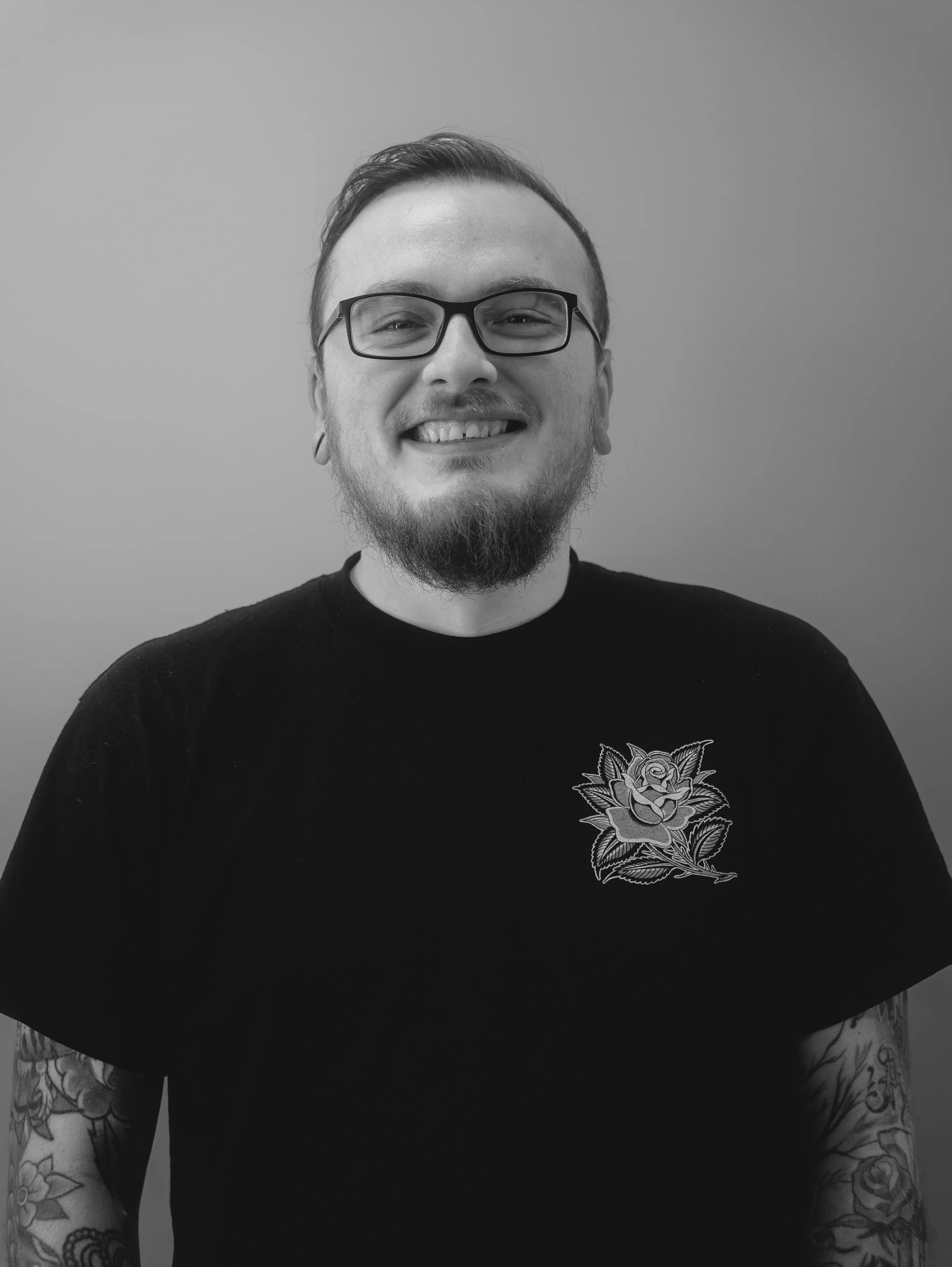 Smiling person with tattoos and gauged ears wearing a black shirt against a gray background.