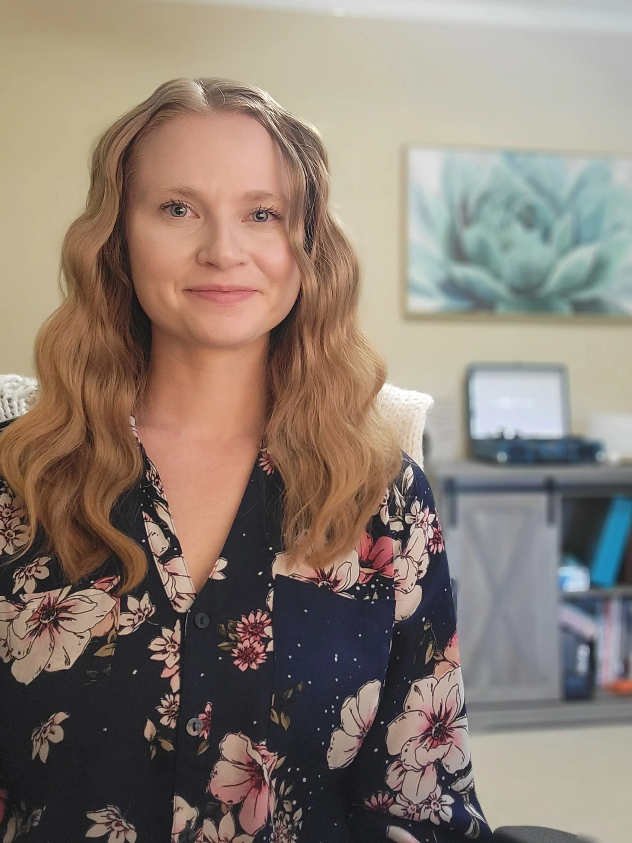 A woman with long, wavy, blonde hair and blue eyes smiling gently, wearing a navy floral blouse, sitting indoors with a blurred background that includes a painting and a work desk with a laptop.