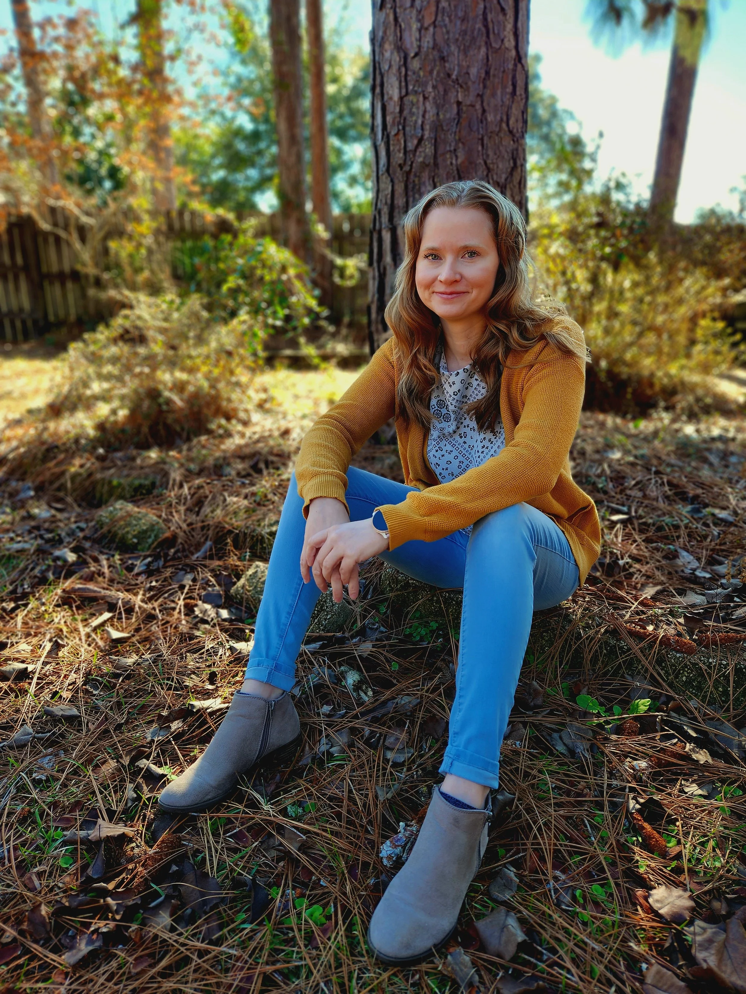 A young woman with wavy brown hair sitting outdoors on a bed of pine needles and fallen leaves, with a background of trees and a wooden fence, smiling at the camera.