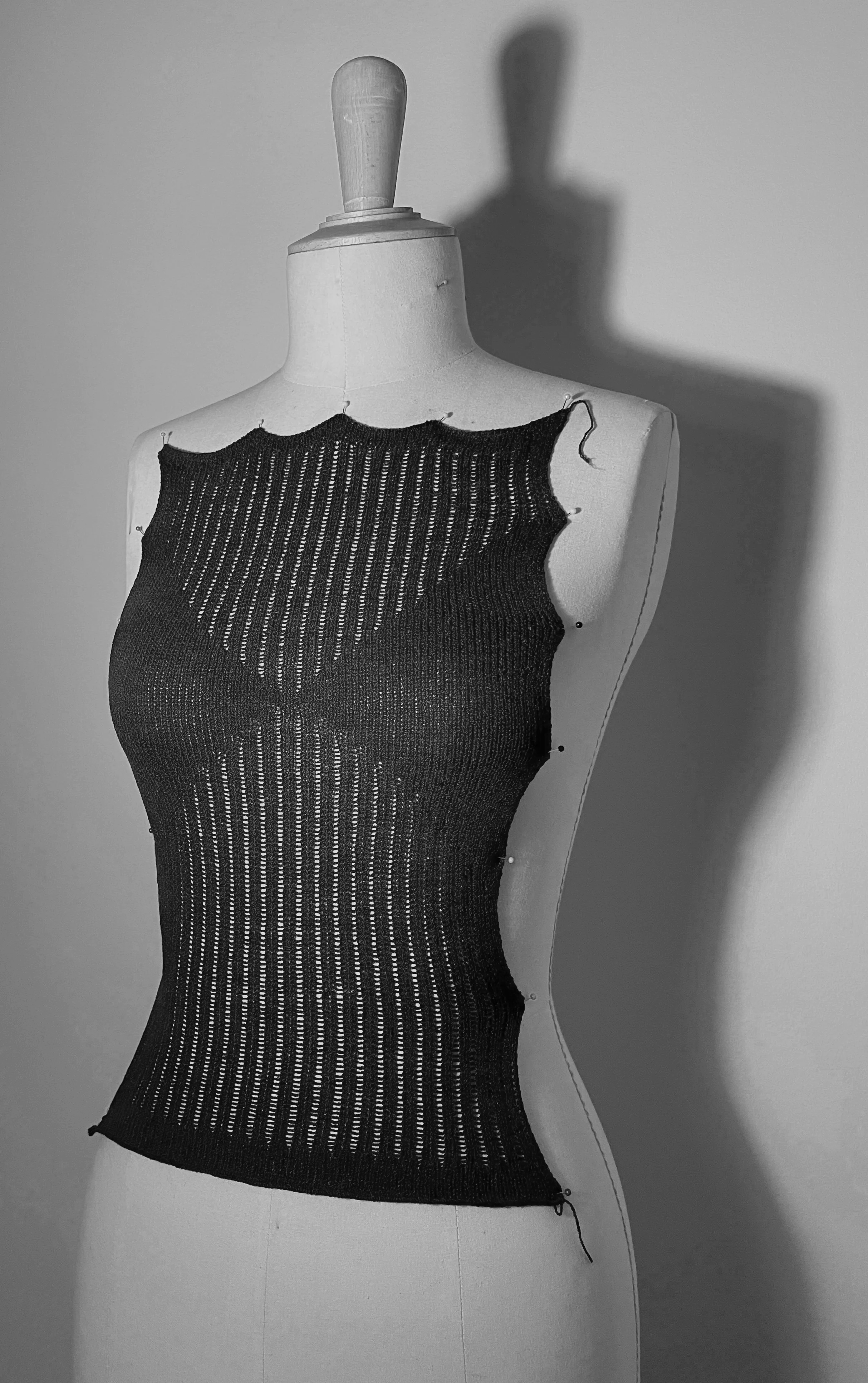 A dress form displays a black, see-through, knitted garment with thin vertical and horizontal stripes, casting a shadow on the wall behind it.