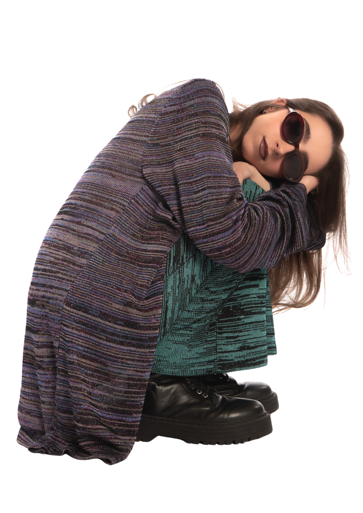 Woman with long brown hair wearing sunglasses, a multicolored textured coat, and black boots, crouching and resting her head on her arms.