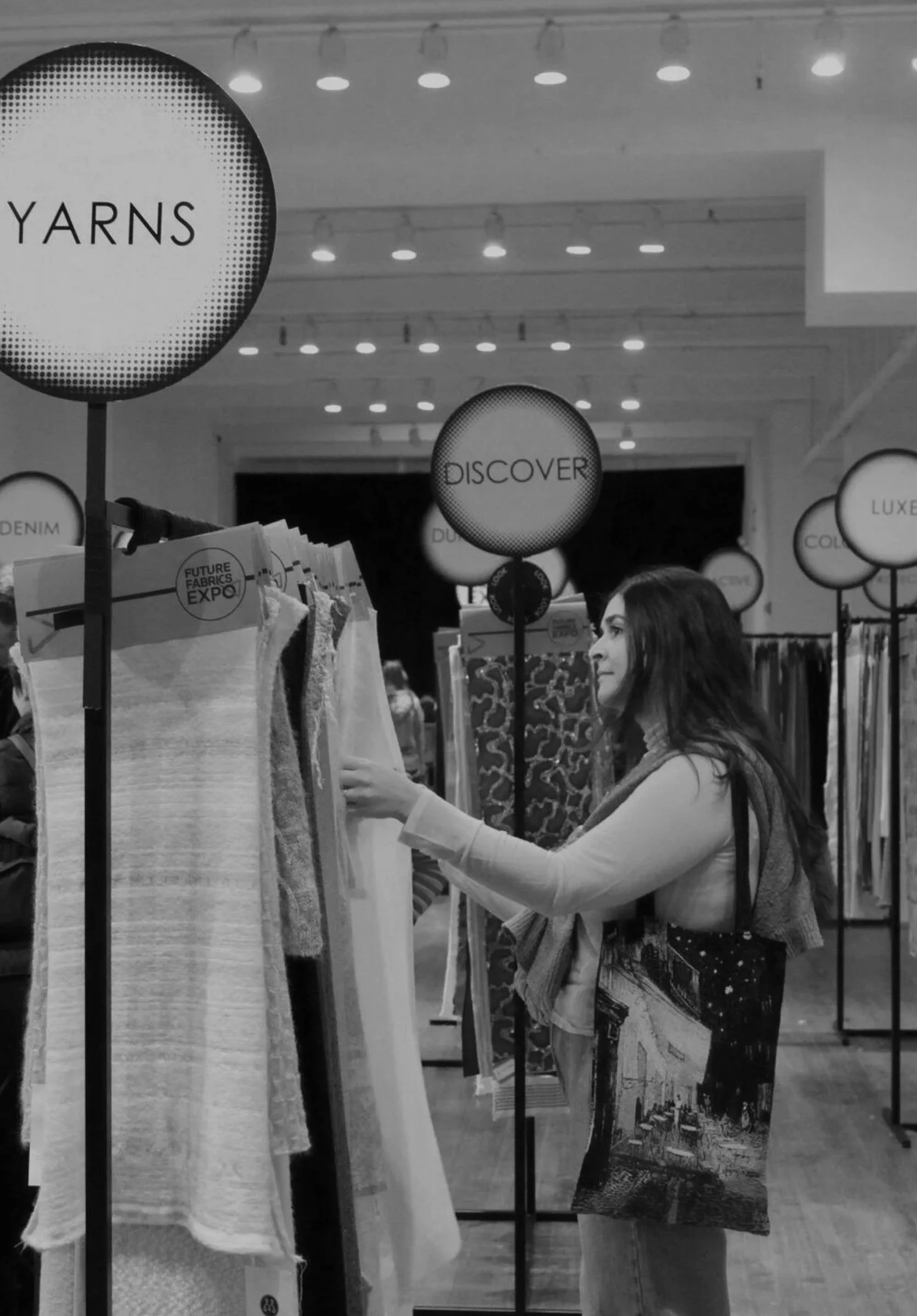 A woman shopping at a fabric store, browsing through different fabric swatches on racks with signage reading 'Discover' and 'Yarns' in the background.