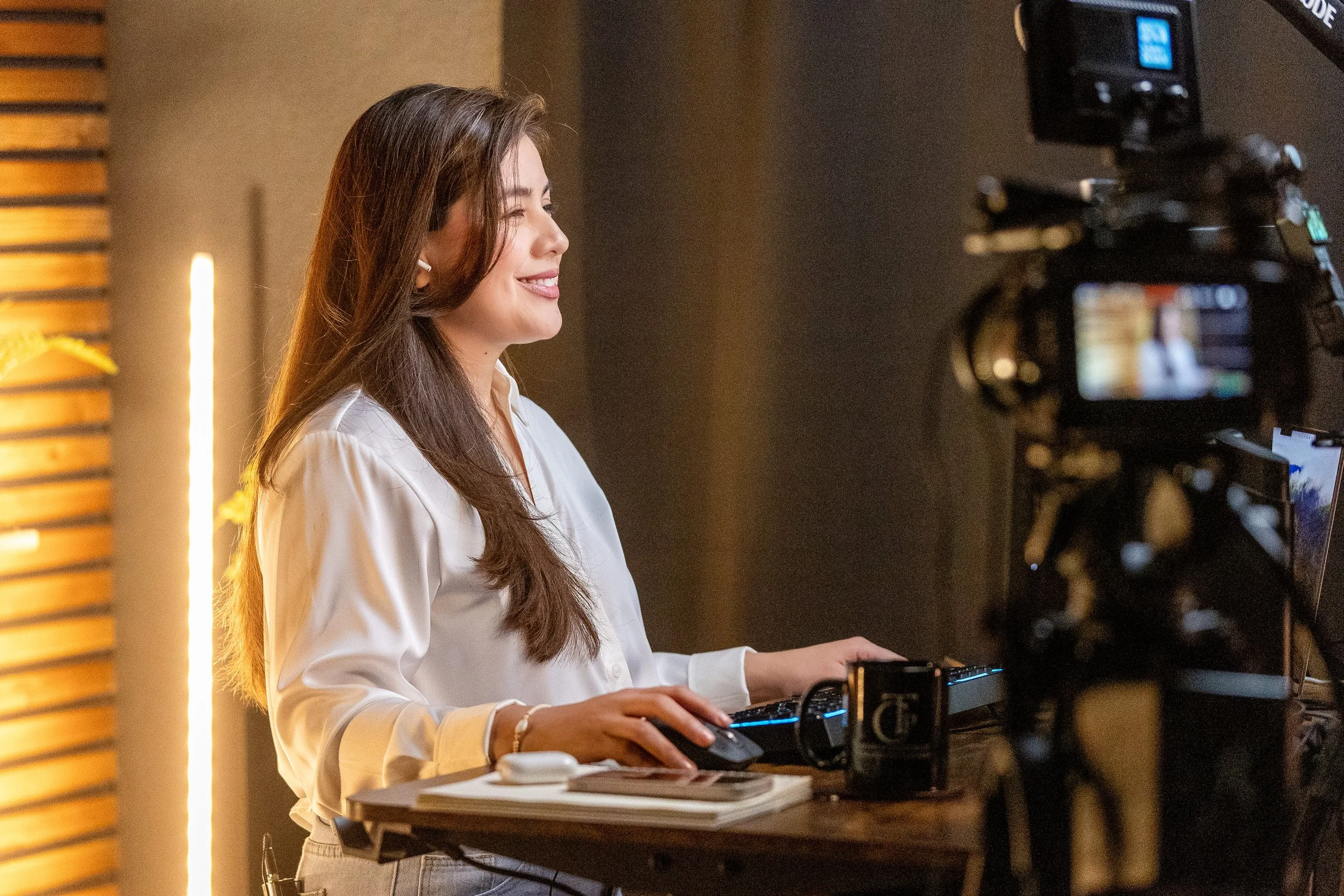 A woman with long brown hair, wearing a white blouse, is sitting at a desk with her right hand on a keyboard and smiling. She is in front of a professional camera pointed at her, with a blurred background featuring a warm lighting setting and a wooden panel on the left.