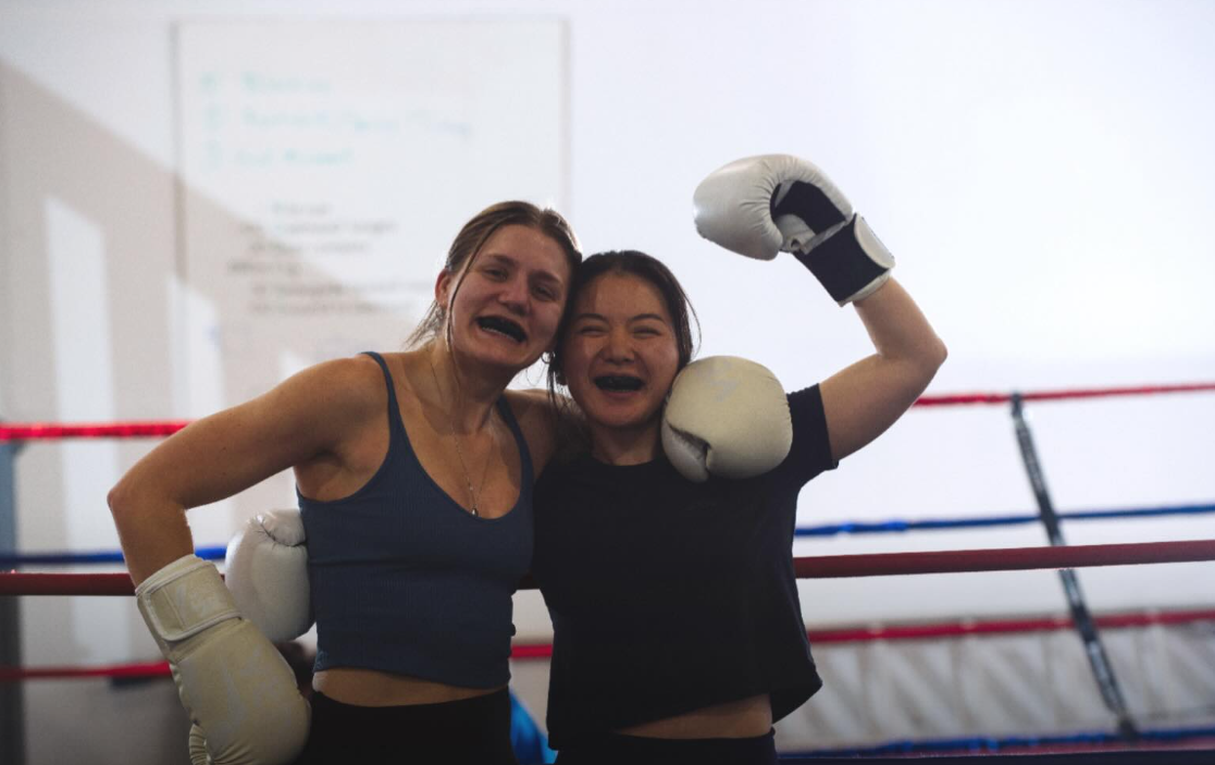 Two women in boxing gloves celebrating inside a boxing ring, one with her arm around the other.