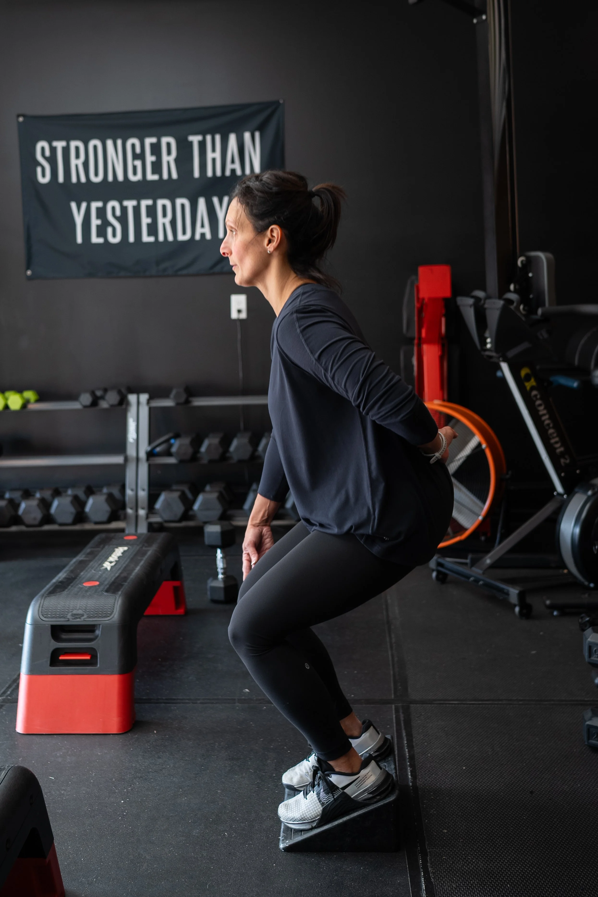 A woman doing a squat exercise on a balance board in a gym. Behind her is a banner reading 'Stronger Than Yesterday' and various gym equipment including dumbbells, a bench, and resistant bands.