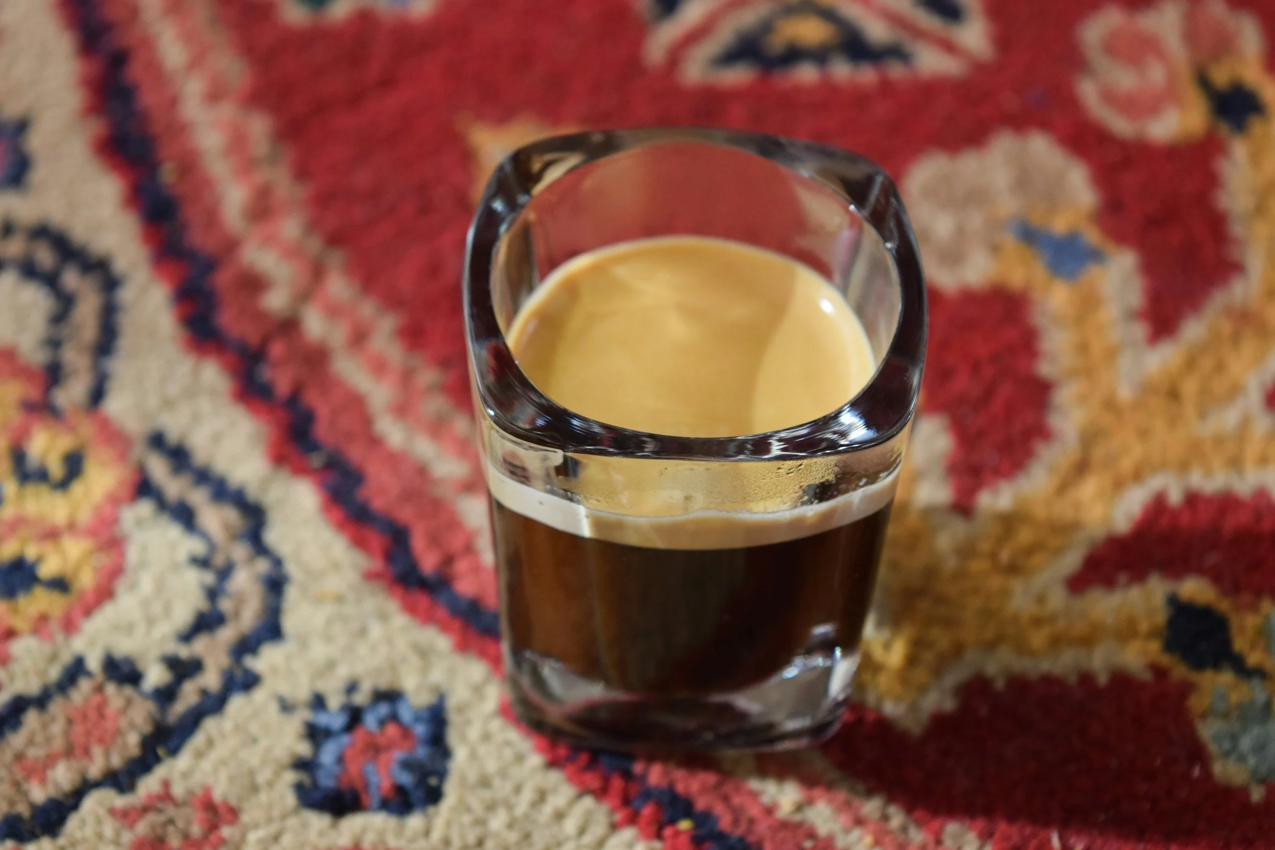 A small drink glass with a brown, coffee colored liquid inside. Glass is on top of Persian style, red, pink, blue, yellow, and white rug.