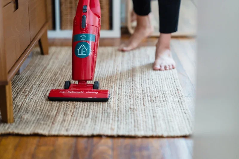 Person vacuuming small rug underneath small wooden storage unit. Vacuum is red and cordless.