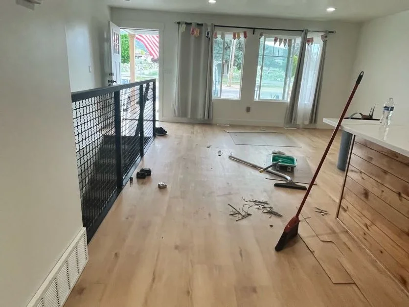 Empty living room with light wood flooring, some cleaning tools and debris on the floor, large window and sliding door with curtains, a railing on the left, and a kitchen counter with a water bottle on the right.