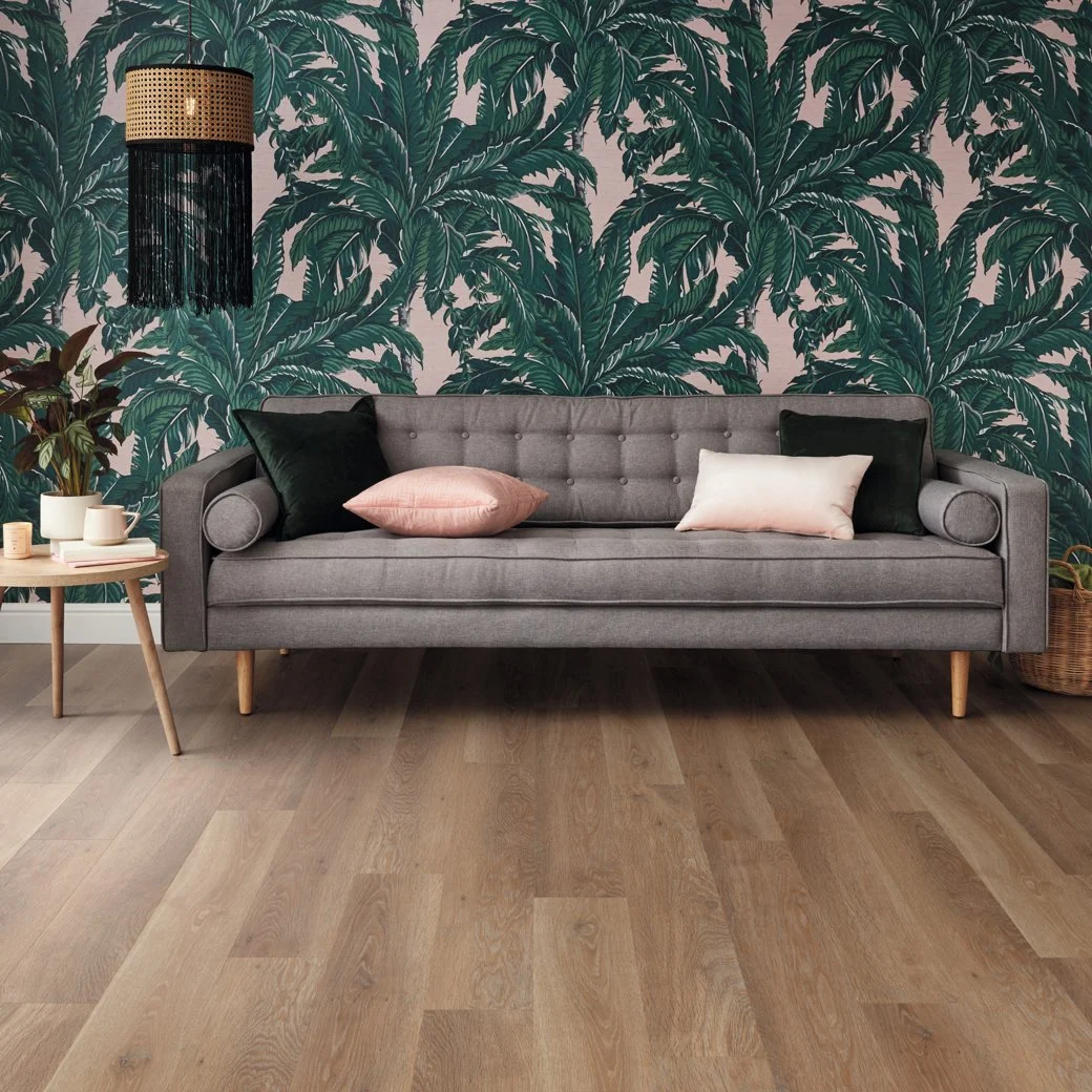 Gray couch with dark green and pale pink pillows, with a busy greenery pattern wallpaper on the back wall. Clicking the image will take you to Karndean's website, who has information on the golden toned floors in the image.