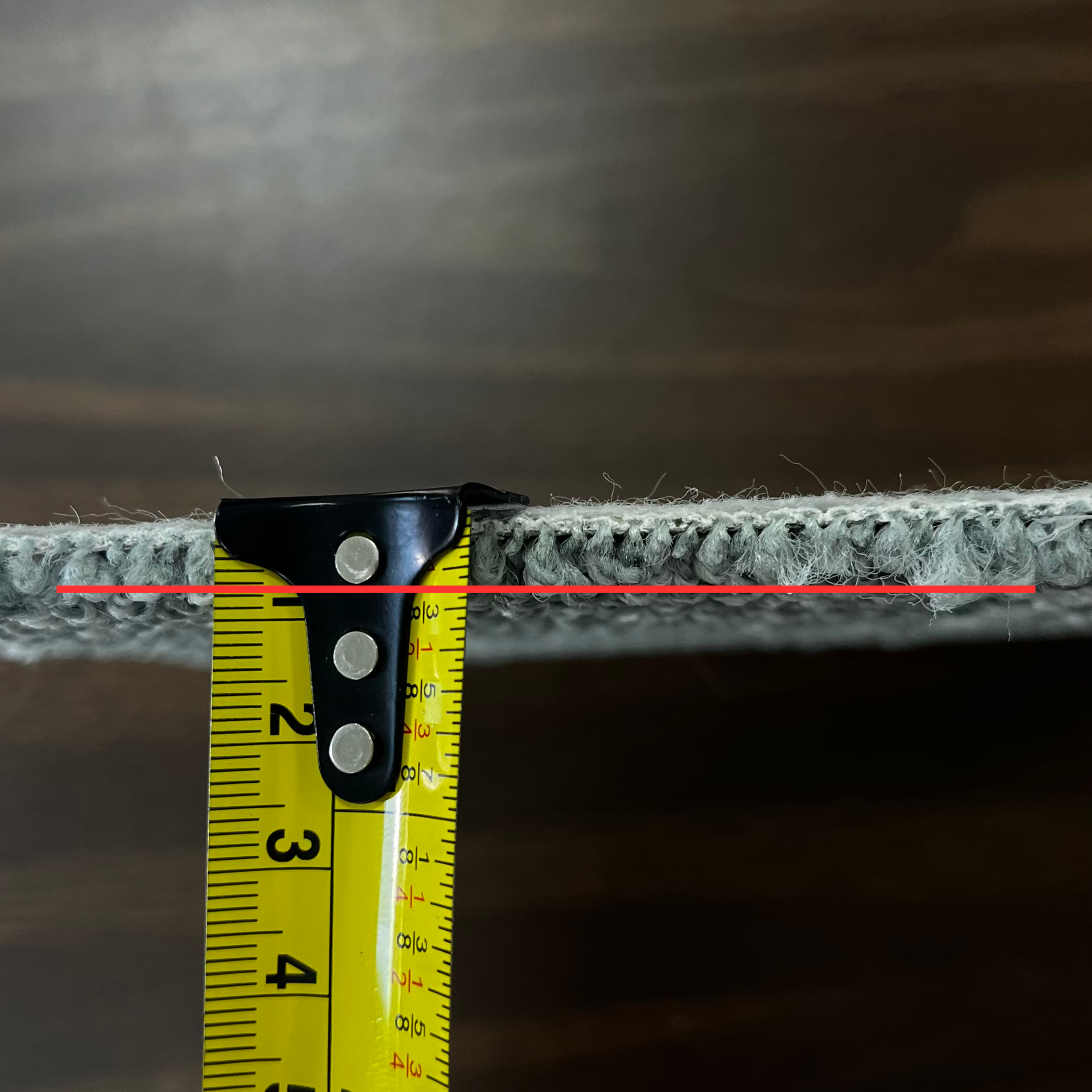 Carpet sample square being measured to see how tall the average pile height is with a red line showing the measurement on a measuring tape