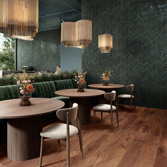 Elegant dining area with round wooden tables, white chairs, green velvet bench, floral centerpieces, green textured wall, and hanging beige chandeliers.