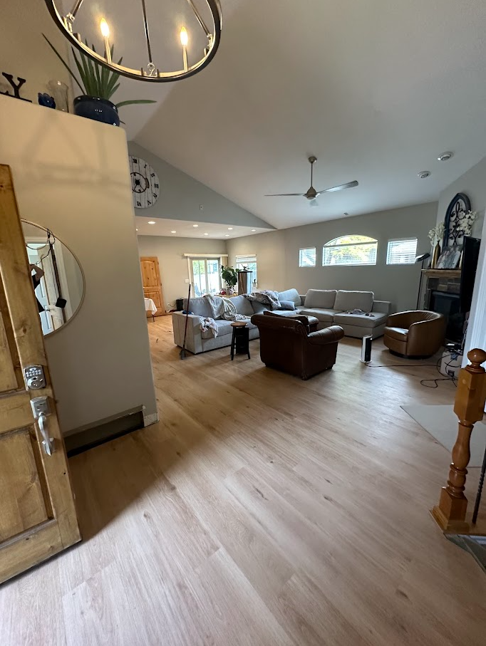 A spacious living room with light wood flooring, multiple sofas, and a ceiling fan. There are large windows letting in natural light, and a fireplace on the right side. Decorative items and plants are visible, with a dining area in the background.