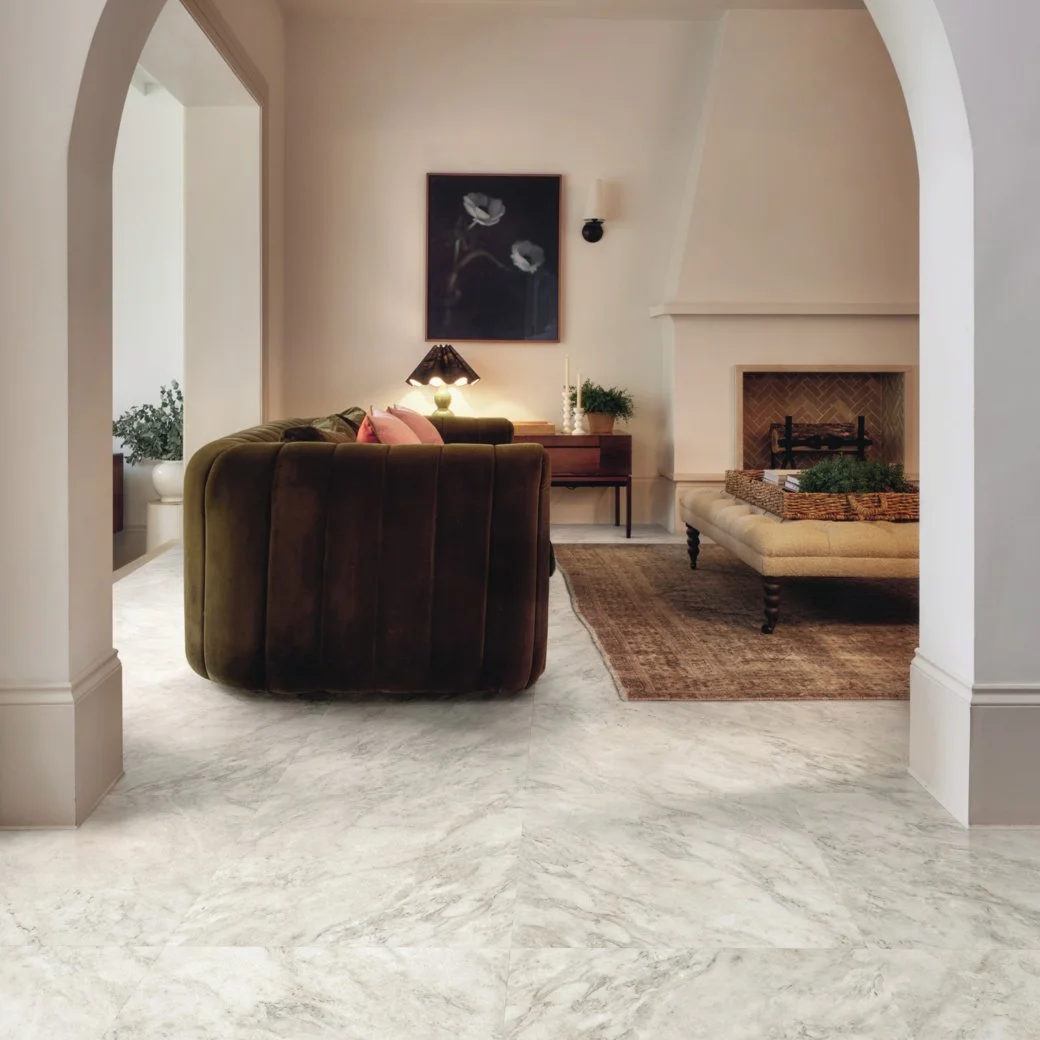Modern living room with marble floors. A green sofa faces into a cloth coffee table. A stucco fireplace with herringbone break inside the hearth takes the back wall. 