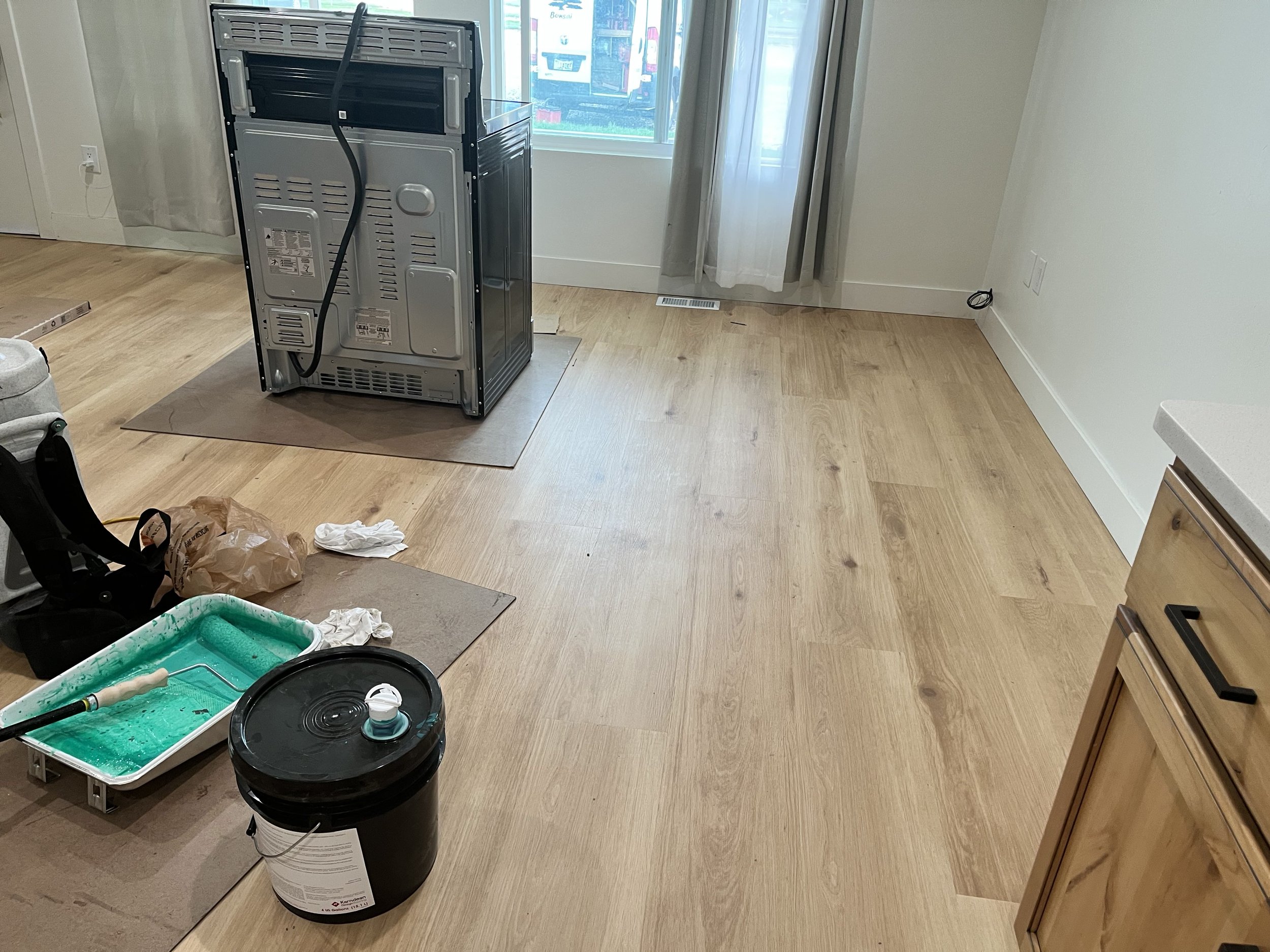 Room with light-colored wooden floors, a window with white curtains, and various objects including a black barrel container, a green and black pet bed, a trash bag, a spray bottle, a black container with a white lid, and dismantled appliances or furniture.