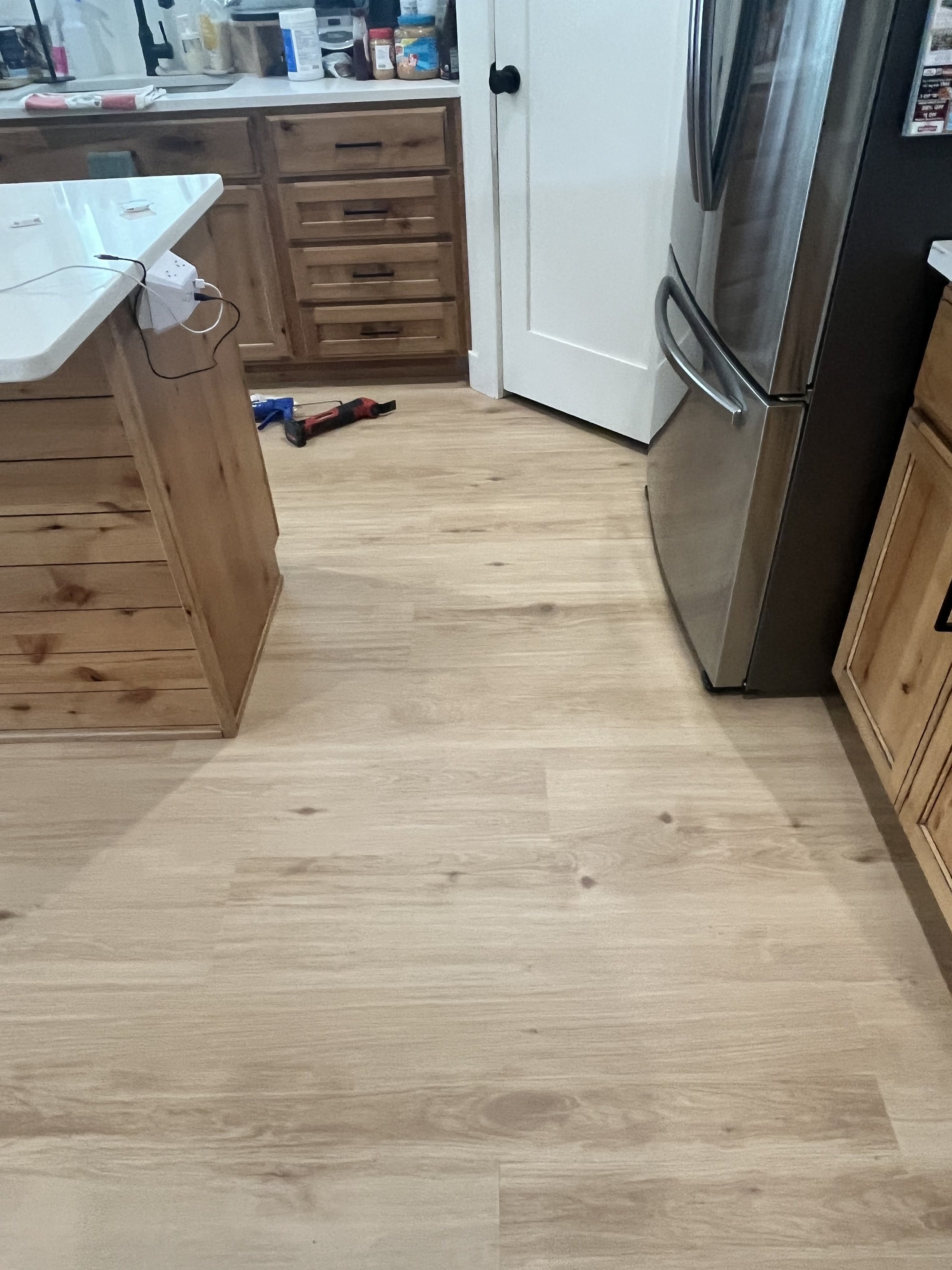 A kitchen with wooden cabinets, a white countertop, stainless steel refrigerator, and a light wood floor. There is a small cordless drill on the floor and some items on the countertop.