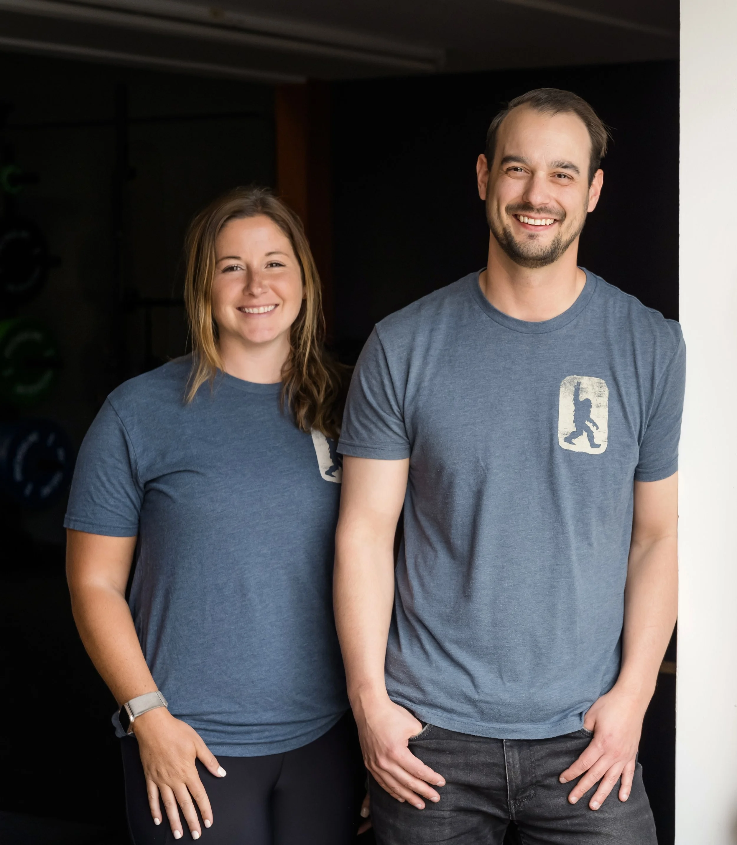 Two smiling people in casual blue T-shirts standing against a black and white background.