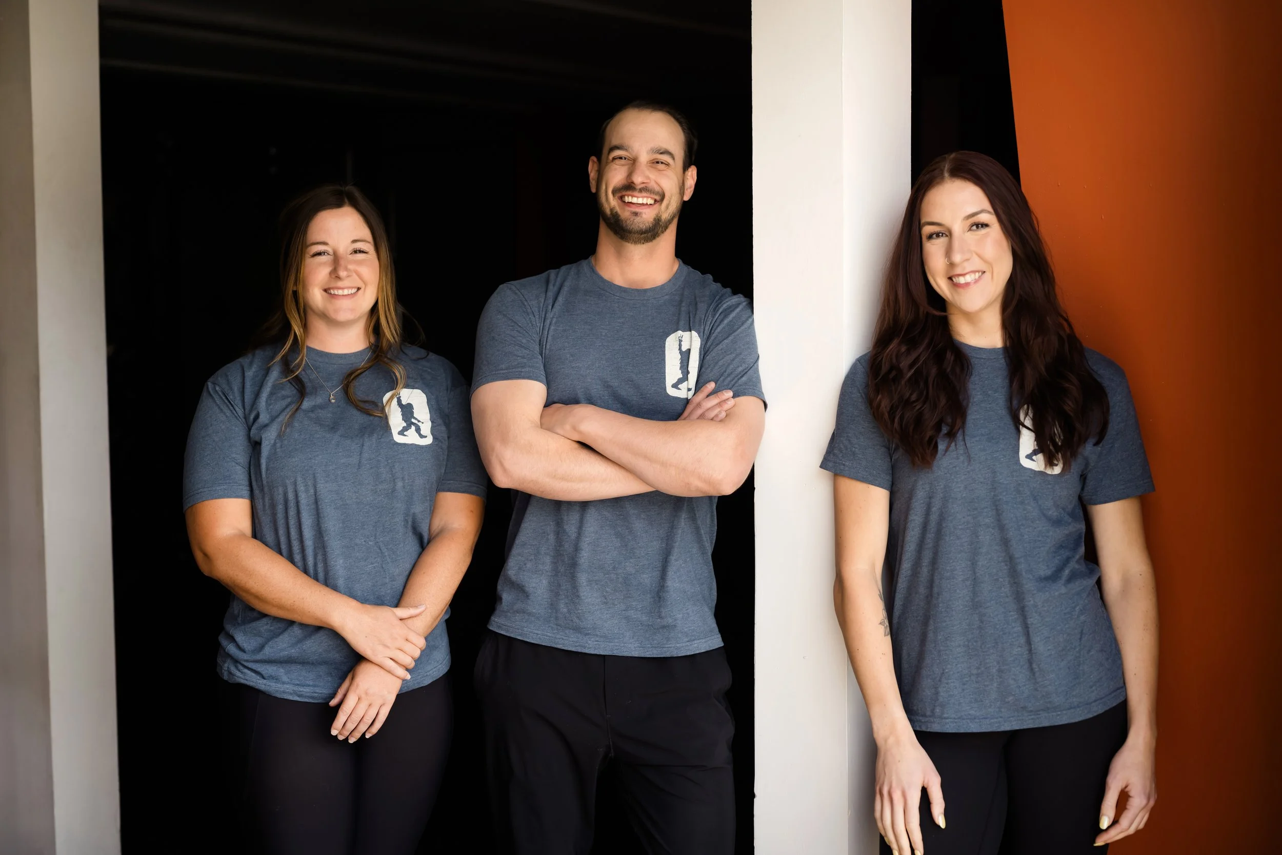 Three smiling people in casual blue T-shirts standing against a black and white background.