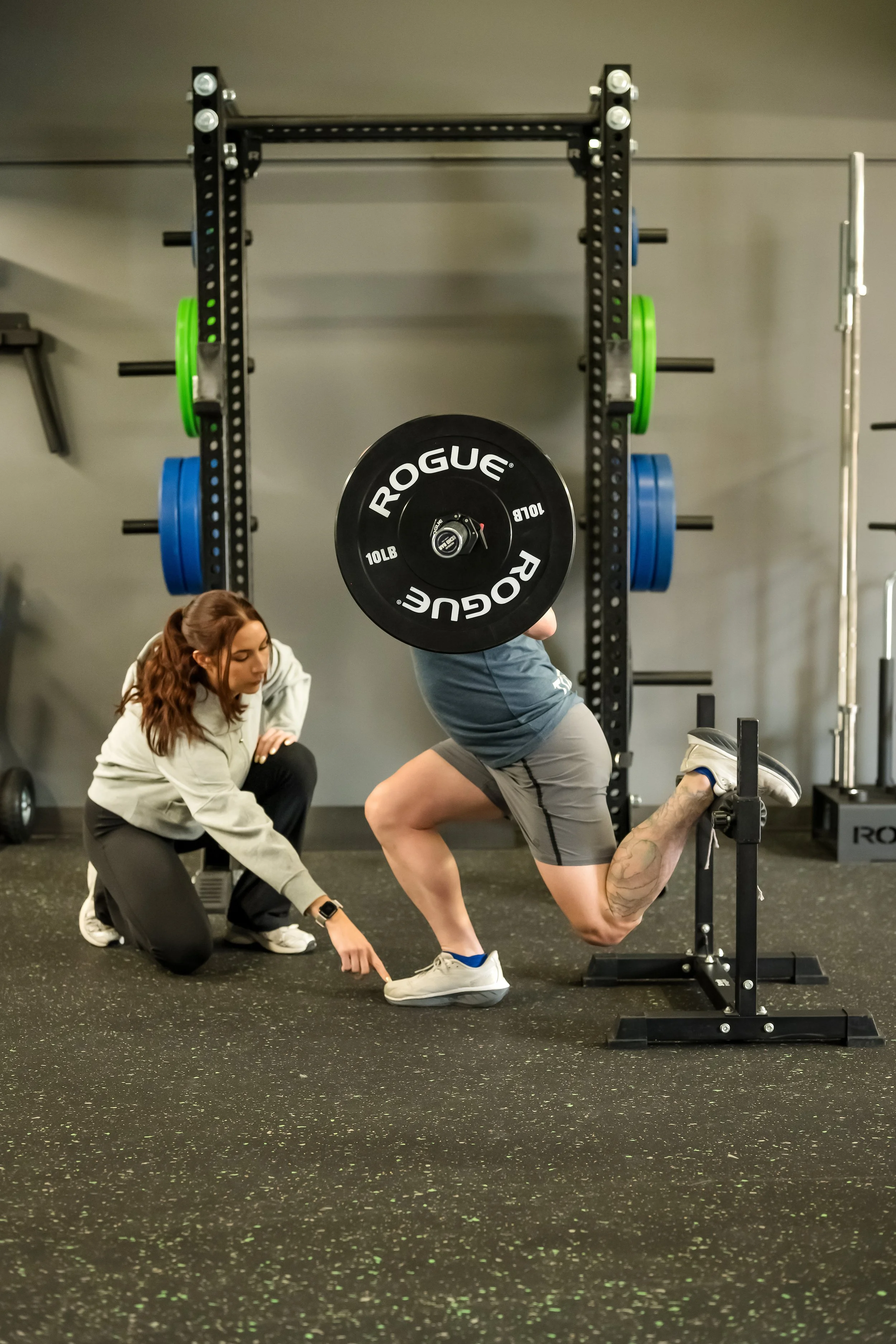 male patient in a blue shirt being assisted by a female physical therapist in a grey sweater with a rack and weights in the background