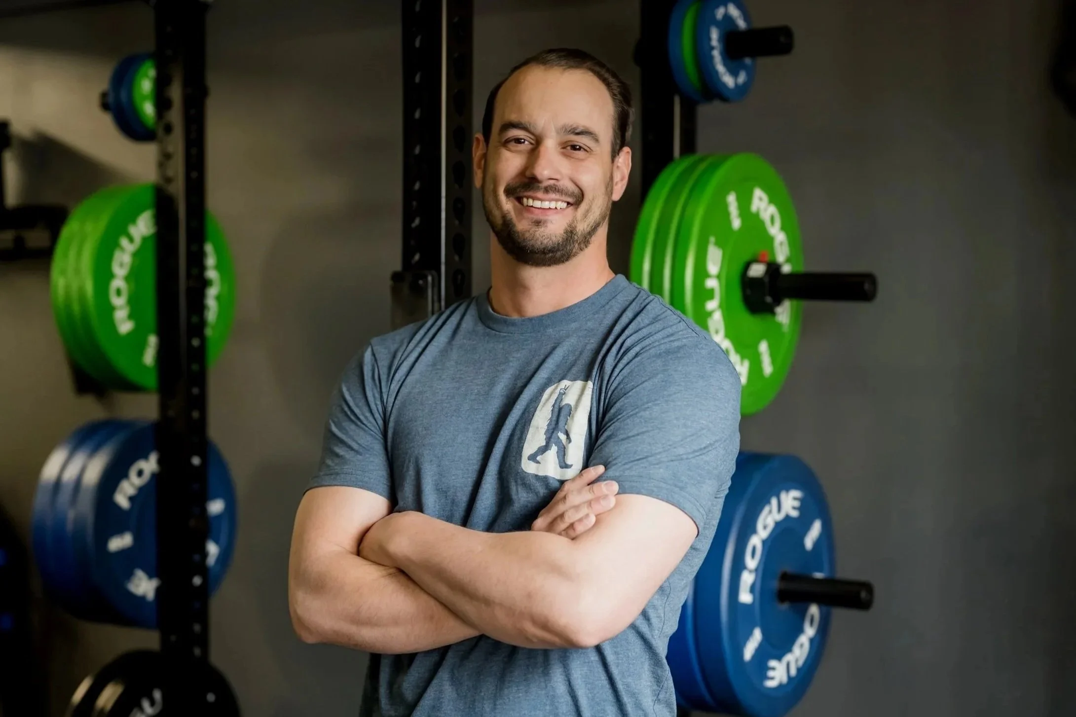 A man with short brown hair, a beard, and a gray t-shirt standing with arms crossed, smiling inside a gym with weights and exercise equipment in the background.