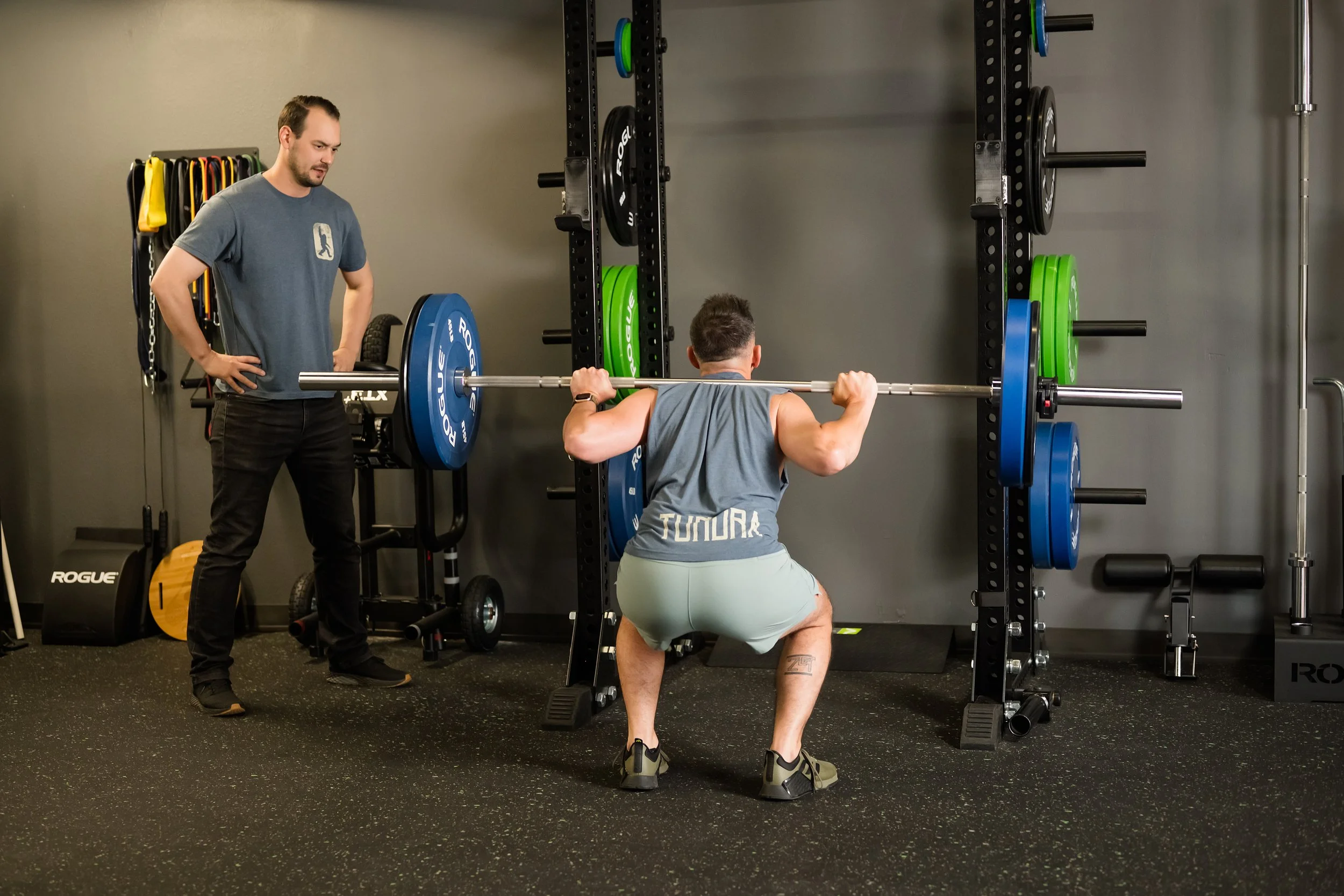 male in a blue tank top squatting being assisted by a male physical therapist in a blue shirt with weights in the background